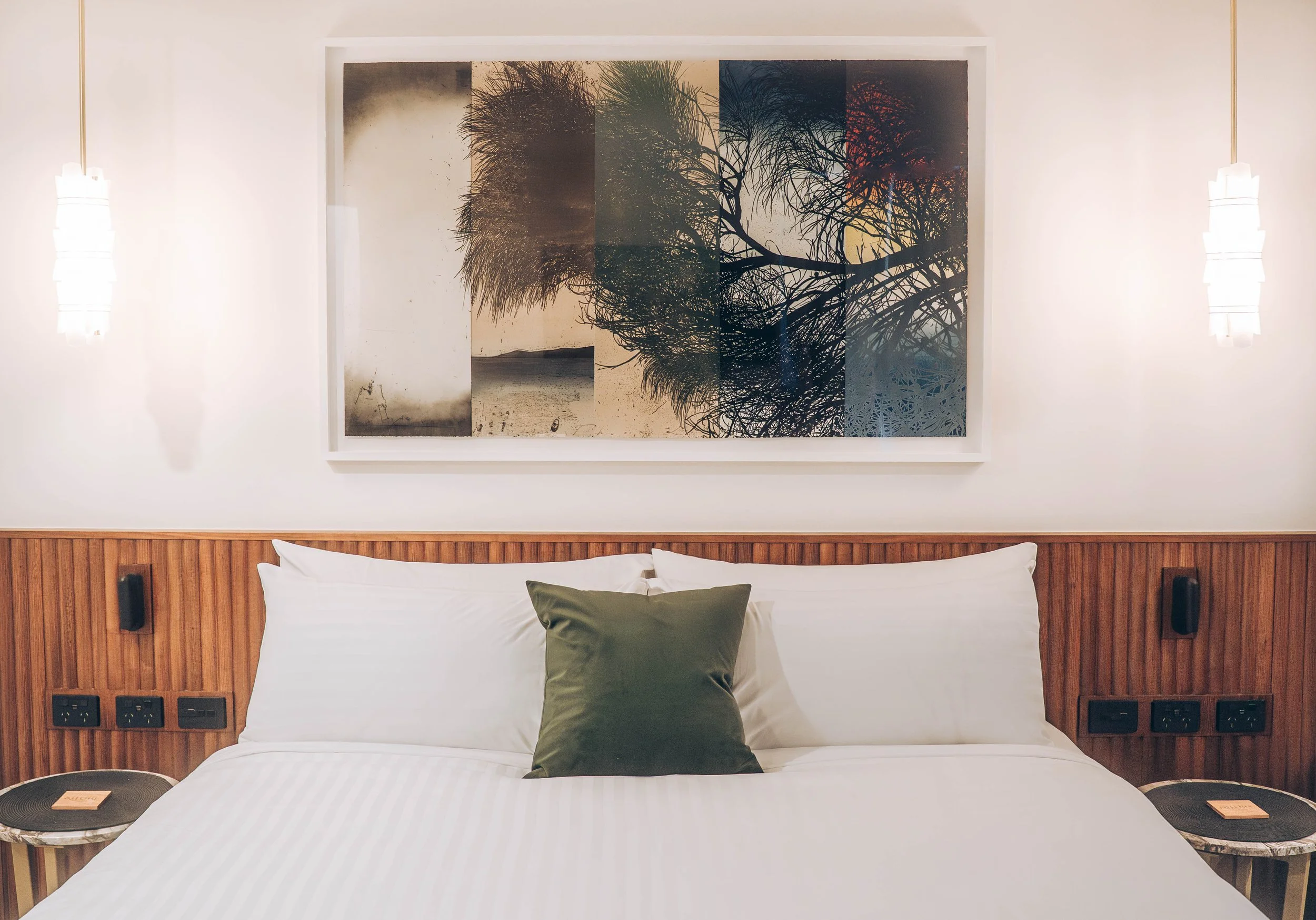 Bedroom interior with a double bed, white linens, green pillow, abstract artwork above the headboard, pendant lights, and wooden accents.