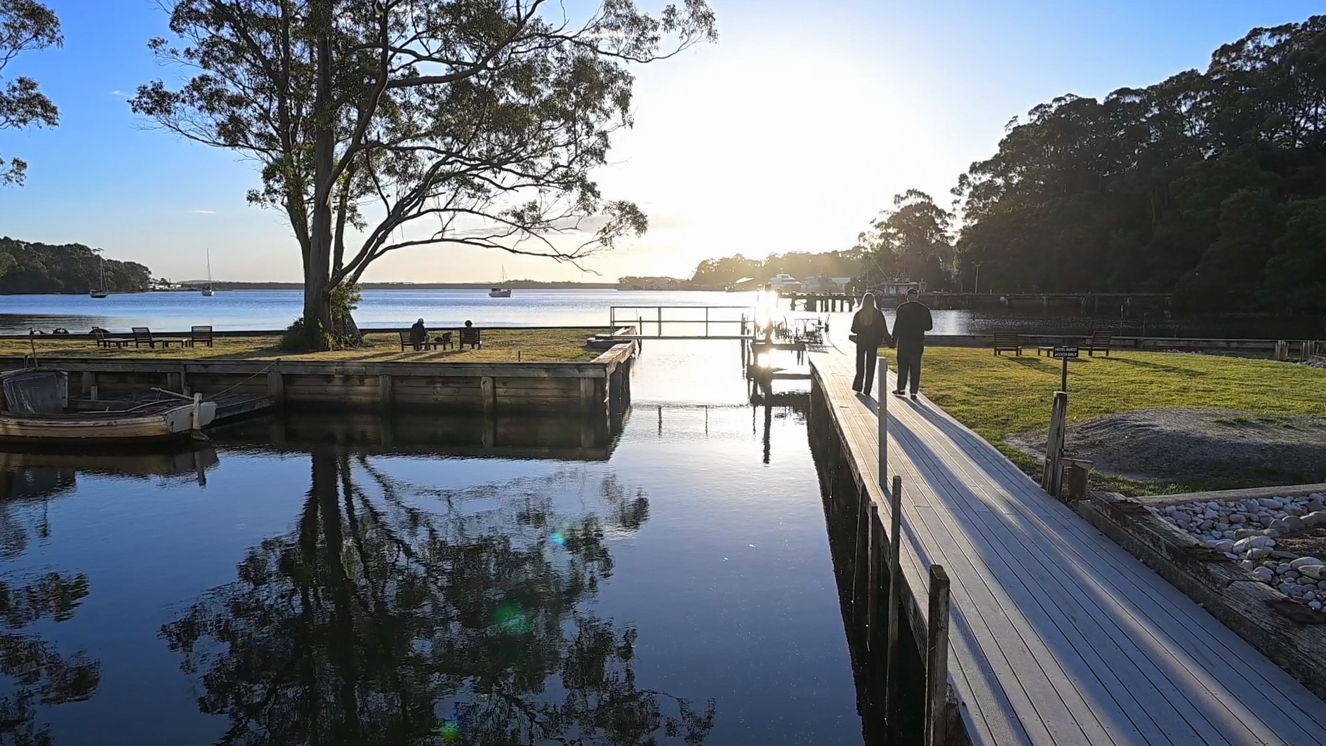 A serene waterfront scene during sunset with two people walking hand in hand along at the Risby Cove marina, a boat moored in the calm water, and other individuals sitting on benches near the shoreline, surrounded by trees and a clear sky.