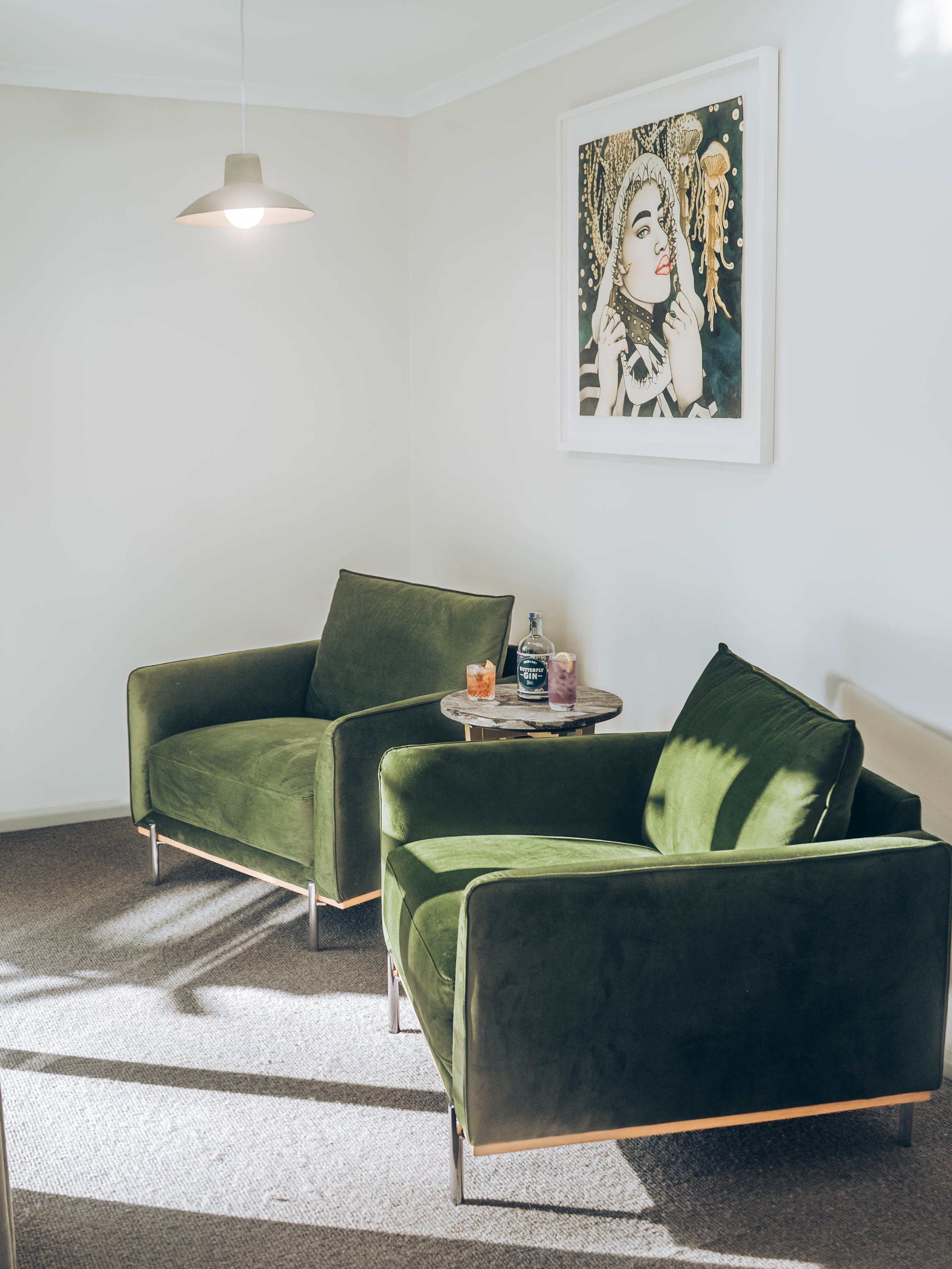 Living room with two green velvet armchairs, a small round wooden side table with drinks and a bottle, a framed artwork of a woman and jellyfish on the white wall, and a hanging ceiling lamp.
