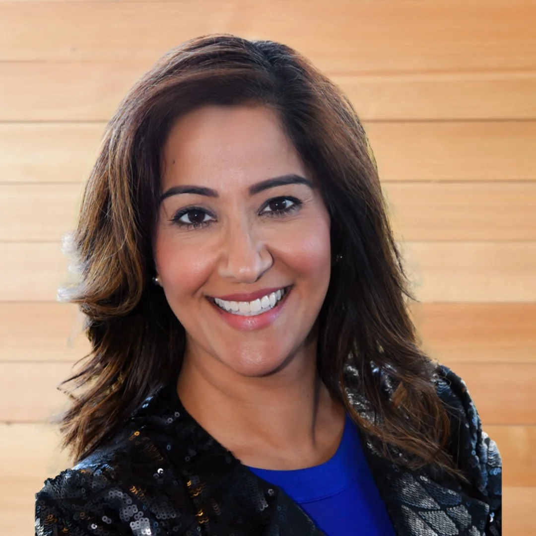 Dr Shimi Kang wearing a sequin jacket and blue top, posing against a wooden background.