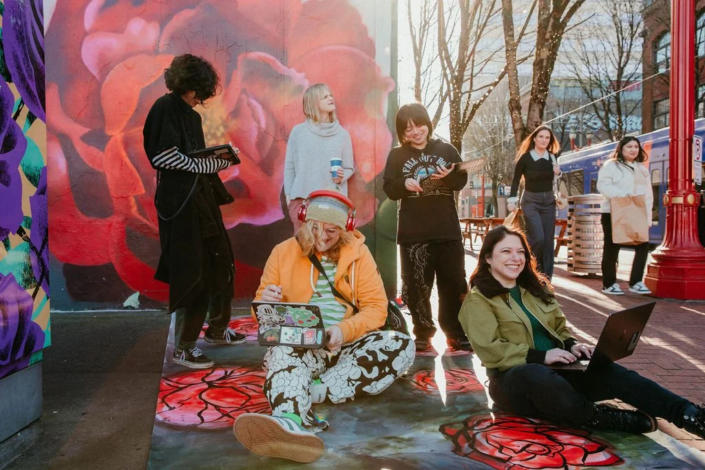 Group of diverse women with laptops and smartphones in an outdoor urban setting near a colorful mural, some sitting and others standing.