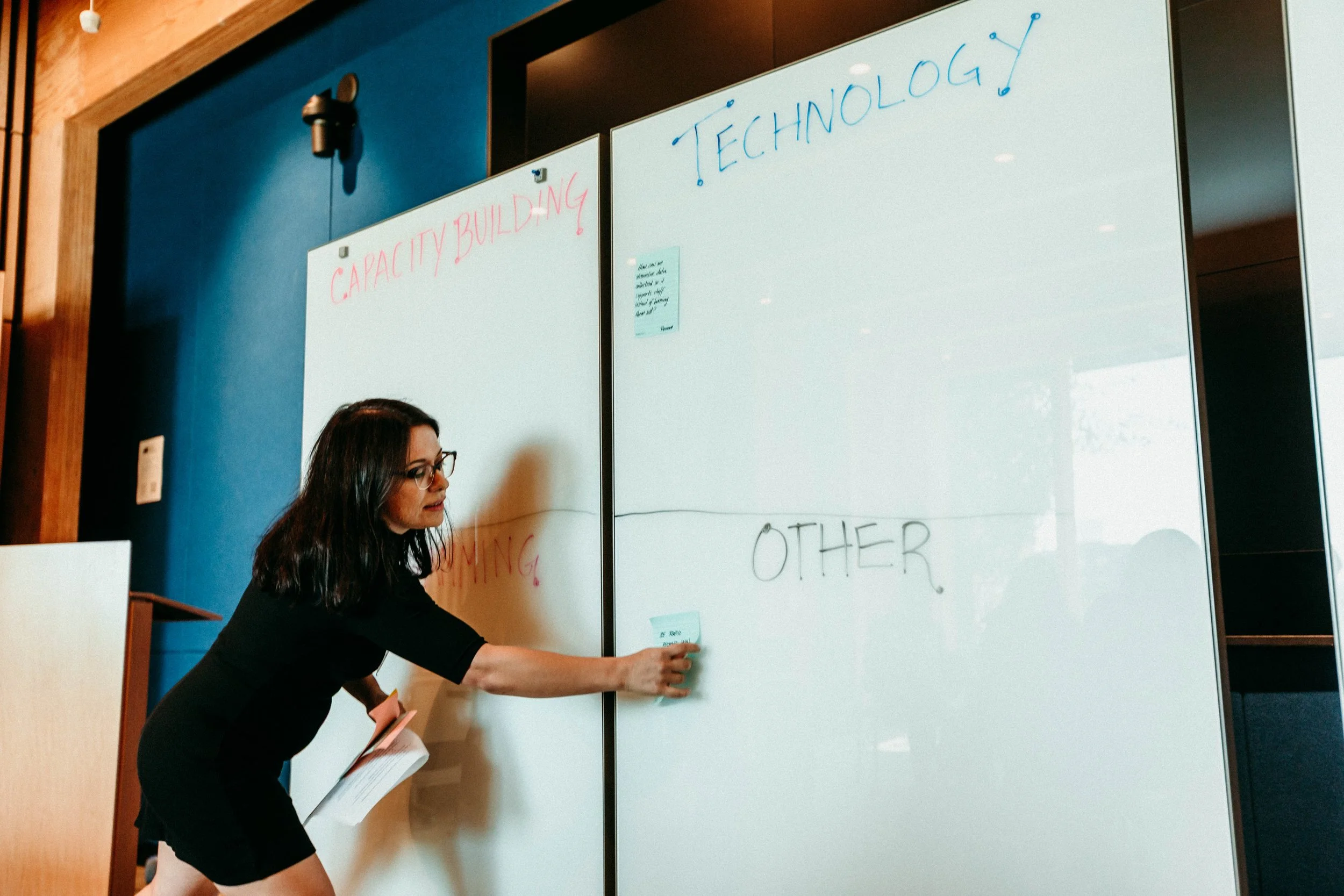 A woman with dark hair and glasses writing on a whiteboard during a presentation or meeting, with text 'CAPACITY BUILDING' and 'TECHNOLOGY' visible on the board.