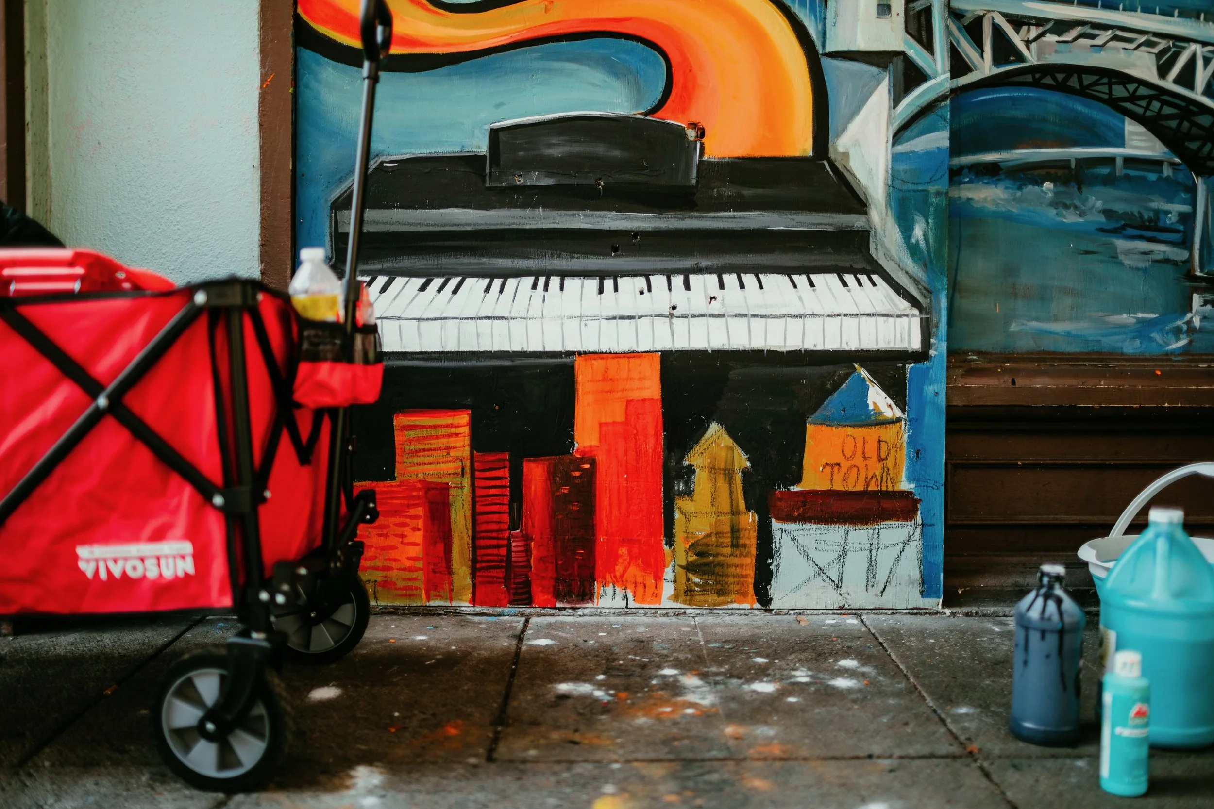 In progress mural of piano with the portland cityscape painted in front. Wagon and painting supplies off to the side