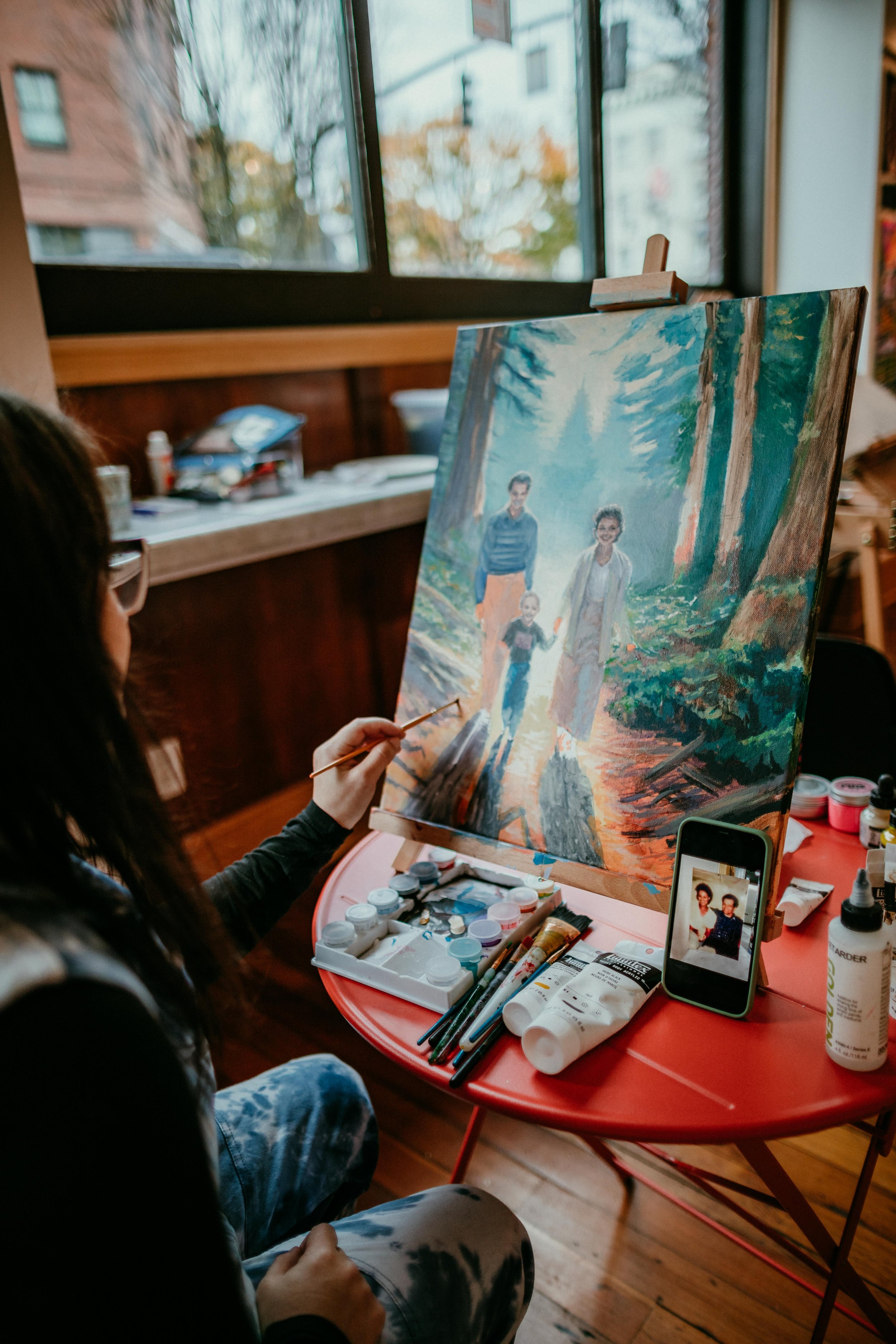 A woman painting a family portrait on a canvas in an art studio with a view of trees outside a window.