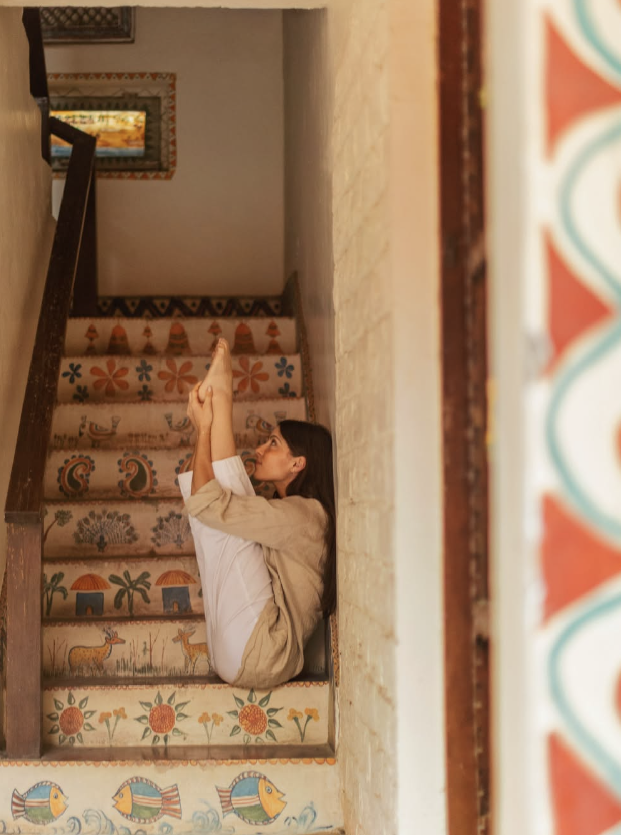 DANA PENSABENE sitting on decorated stairs with colorful patterns in RISHIKESH INDIA DOING A YOGA POSE, holding her hands together in a prayer-like pose. She is indoors with a beige wall on her right and a colorful wall painting in the background.