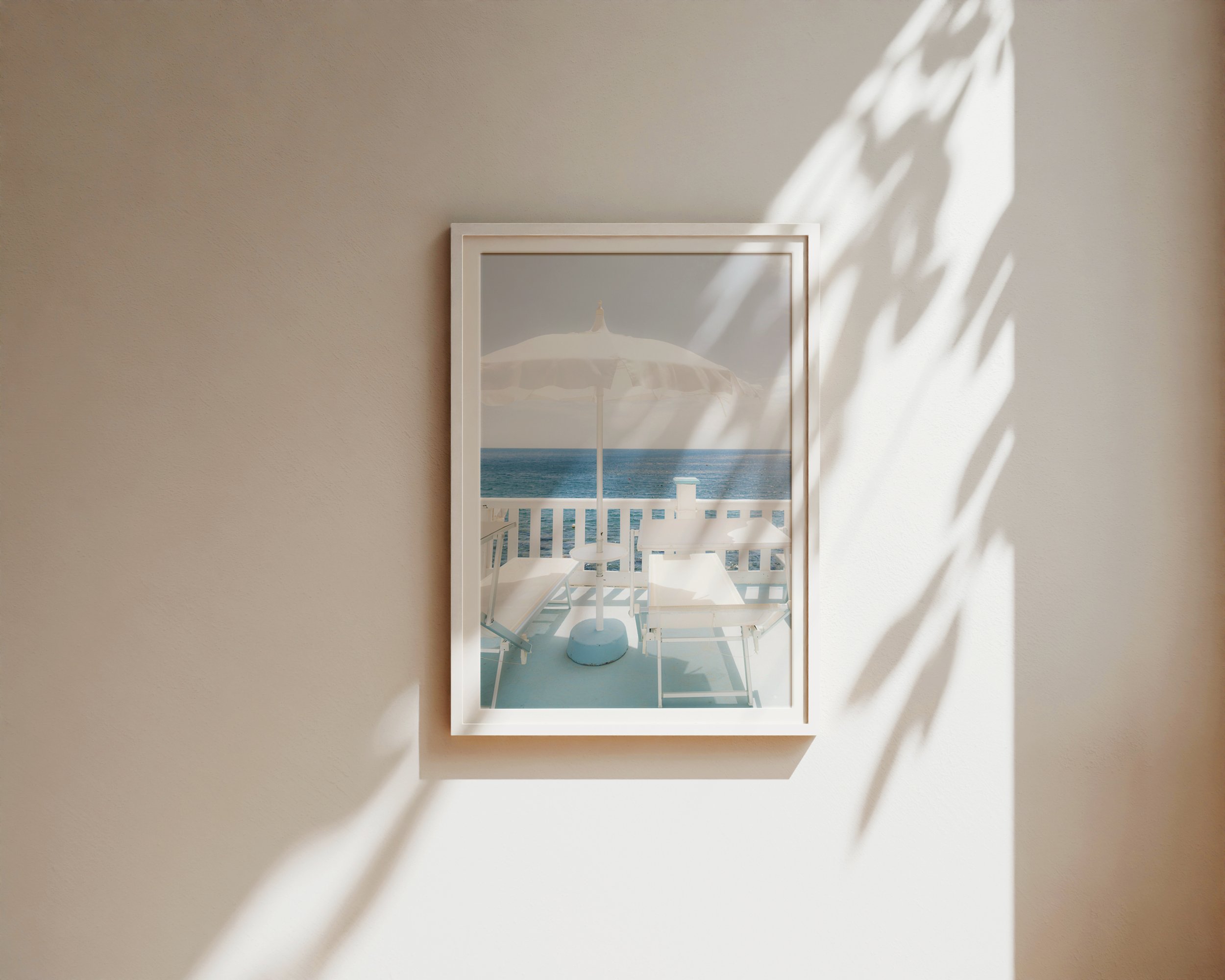 A serene coastal photograph featuring two white lounge chairs and a white umbrella on a pastel-blue balcony overlooking the ocean.