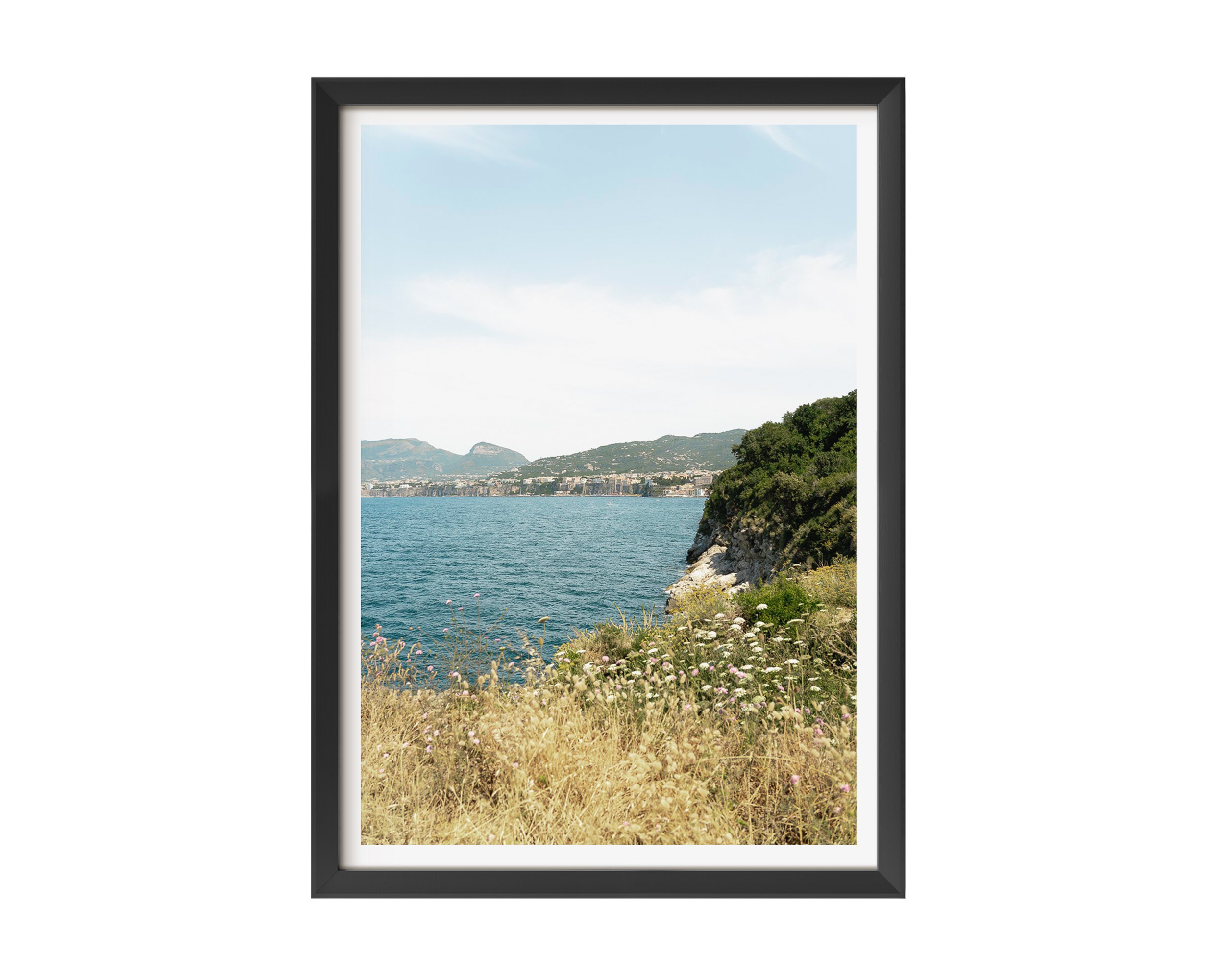 A fine art photograph capturing the quiet beauty of a sunlit coastline. Golden wild grasses and delicate white wildflowers frame a rugged cliff that descends into the calm, deep blue sea.