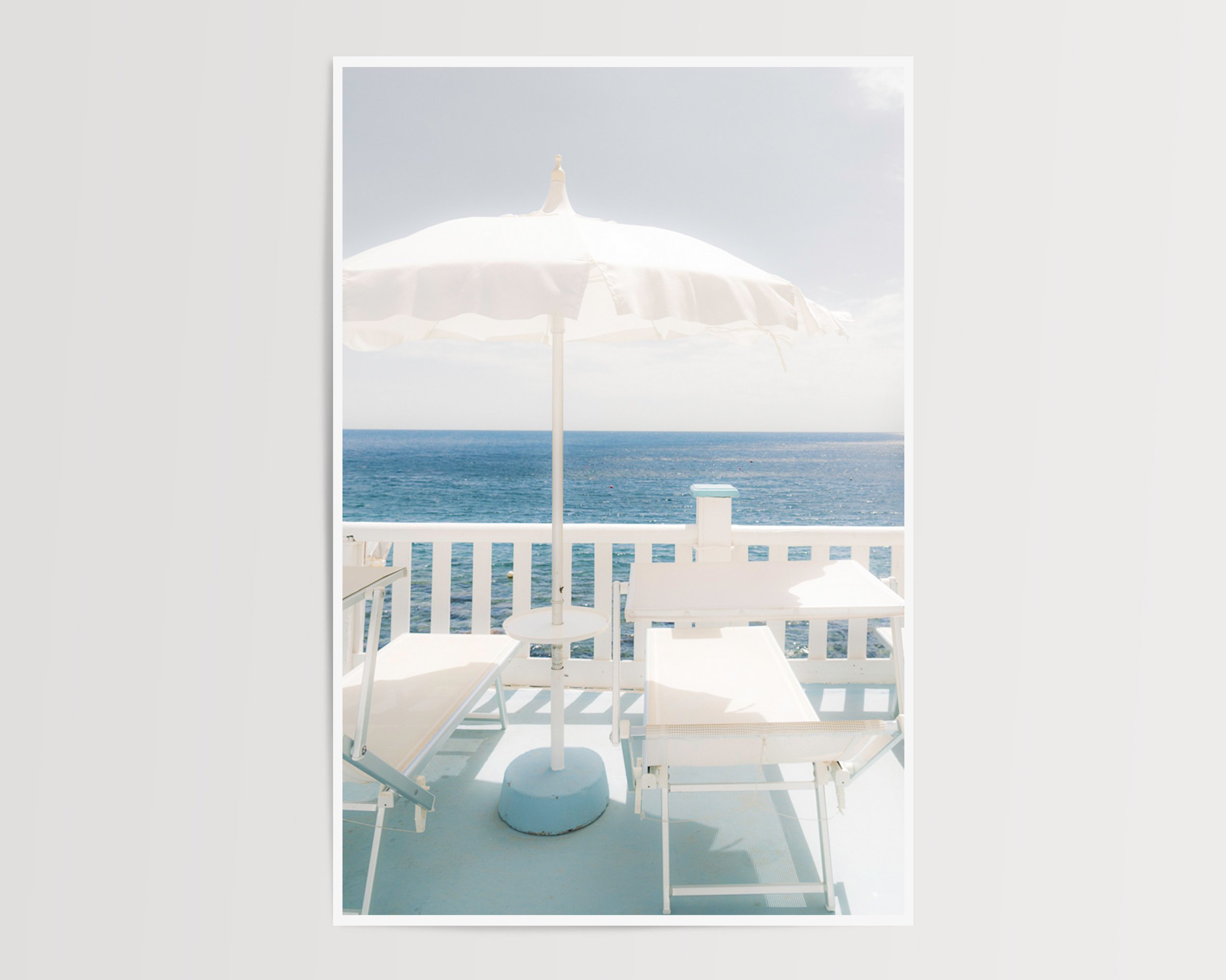 A serene coastal photograph featuring two white lounge chairs and a white umbrella on a pastel-blue balcony overlooking the ocean.