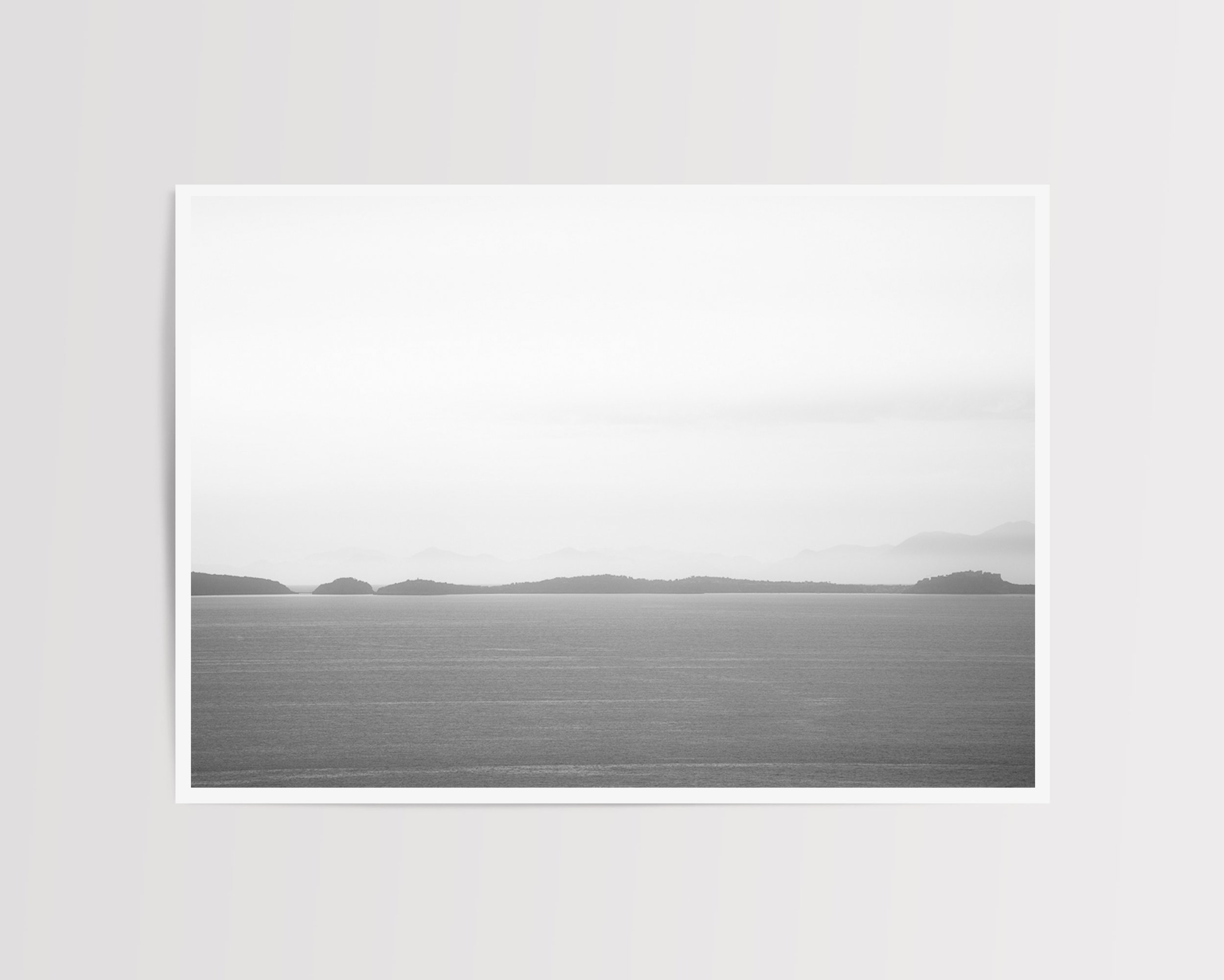 A tranquil black and white fine art photograph capturing a calm sea and misty distant islands beneath a soft, tonal sky.
