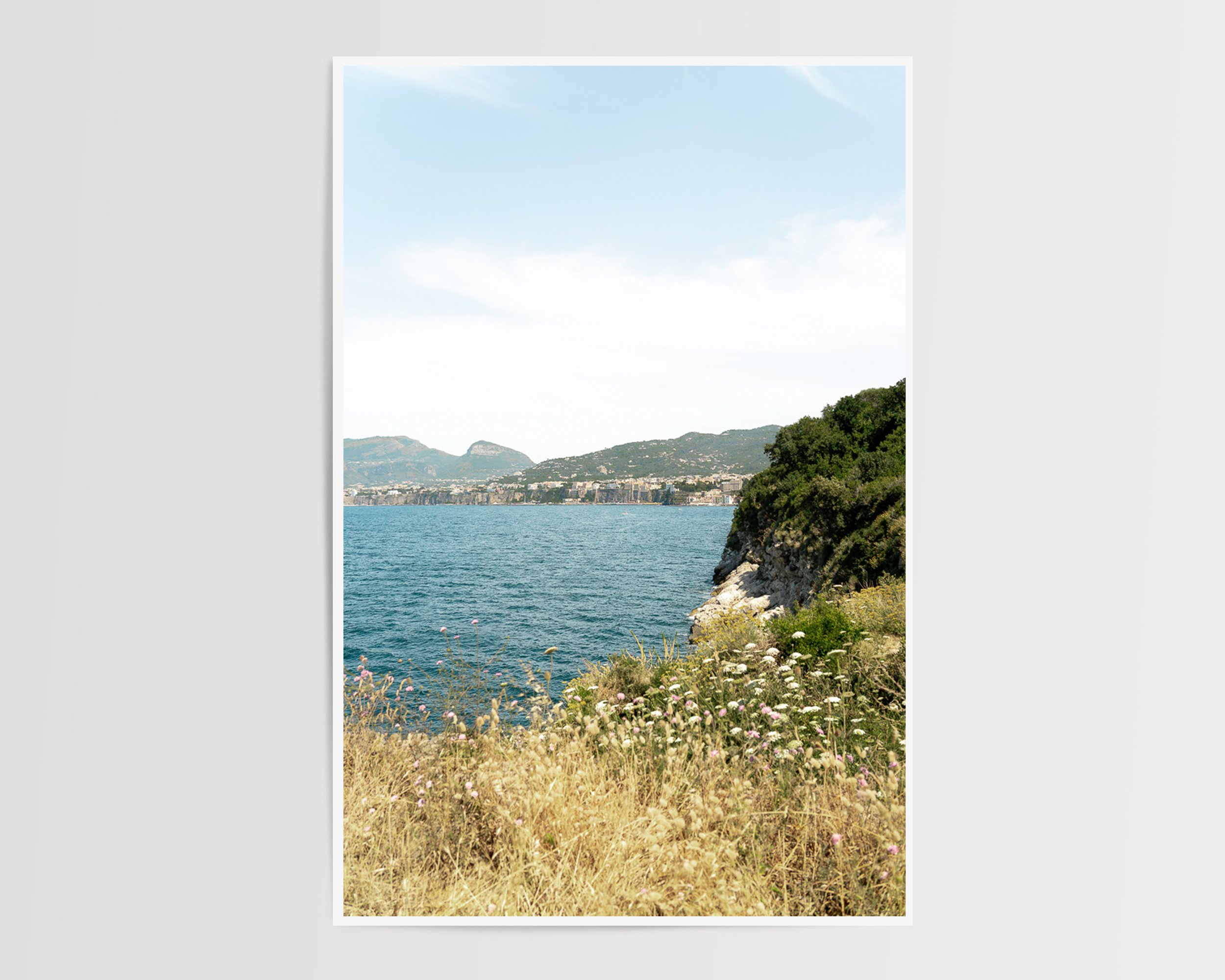 A fine art photograph capturing the quiet beauty of a sunlit coastline. Golden wild grasses and delicate white wildflowers frame a rugged cliff that descends into the calm, deep blue sea.