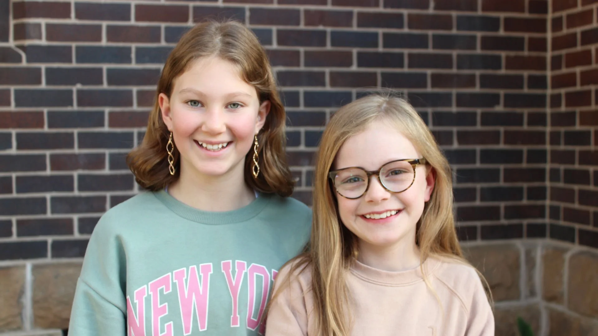 Two teenage girls smiling and standing outdoors in front of a brick wall.