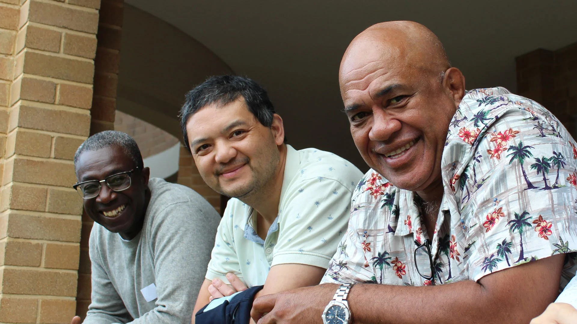 Three men sitting outdoors near a brick wall, smiling at the camera.