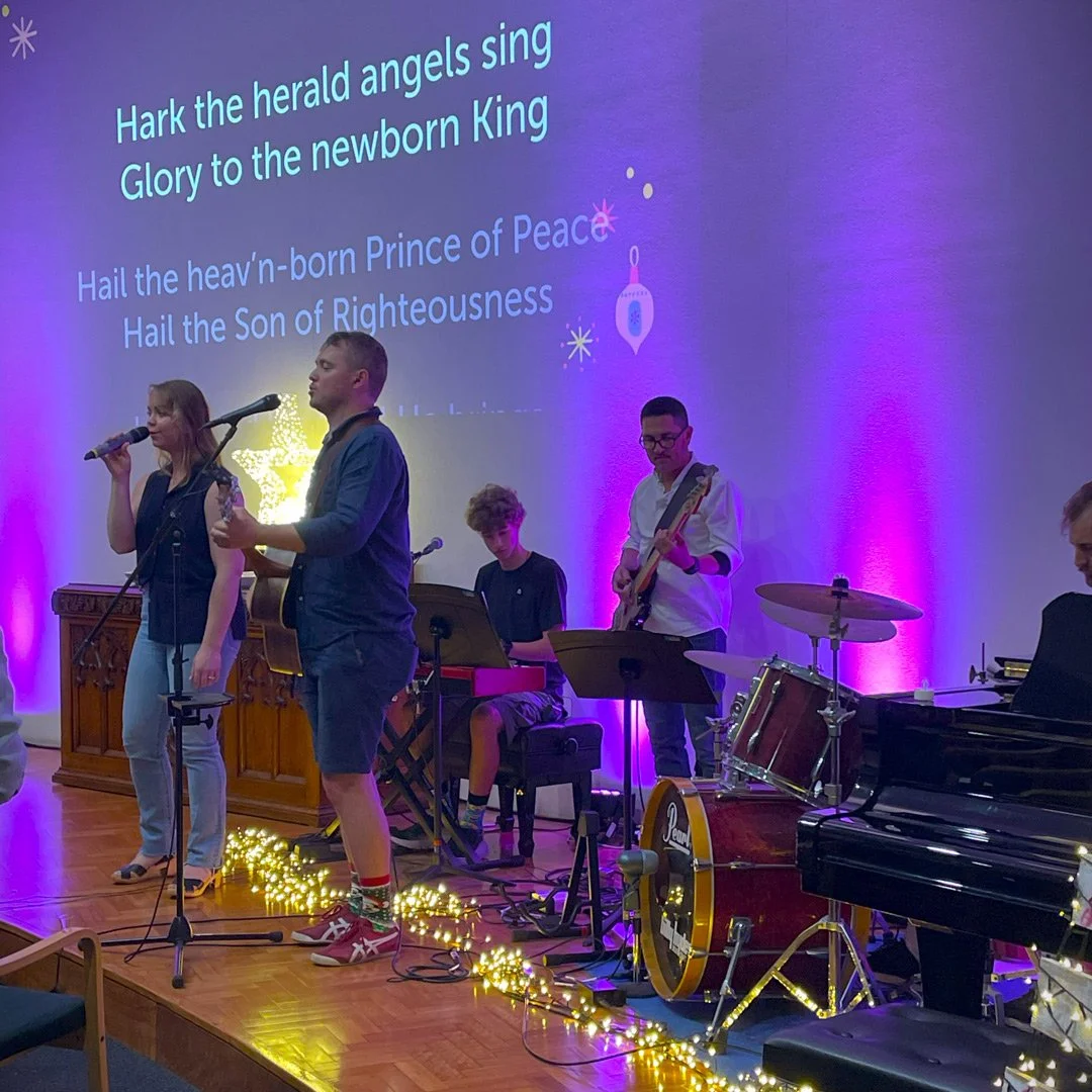 A group of five musicians performing on stage, singing and playing instruments, with a Christmas-themed background and lyrics projected on the wall behind them.