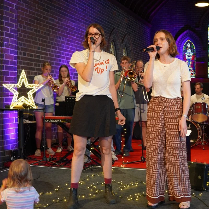 Two women singing into microphones on stage, with a band playing instruments behind them, in a venue with stained glass windows and purple lighting.