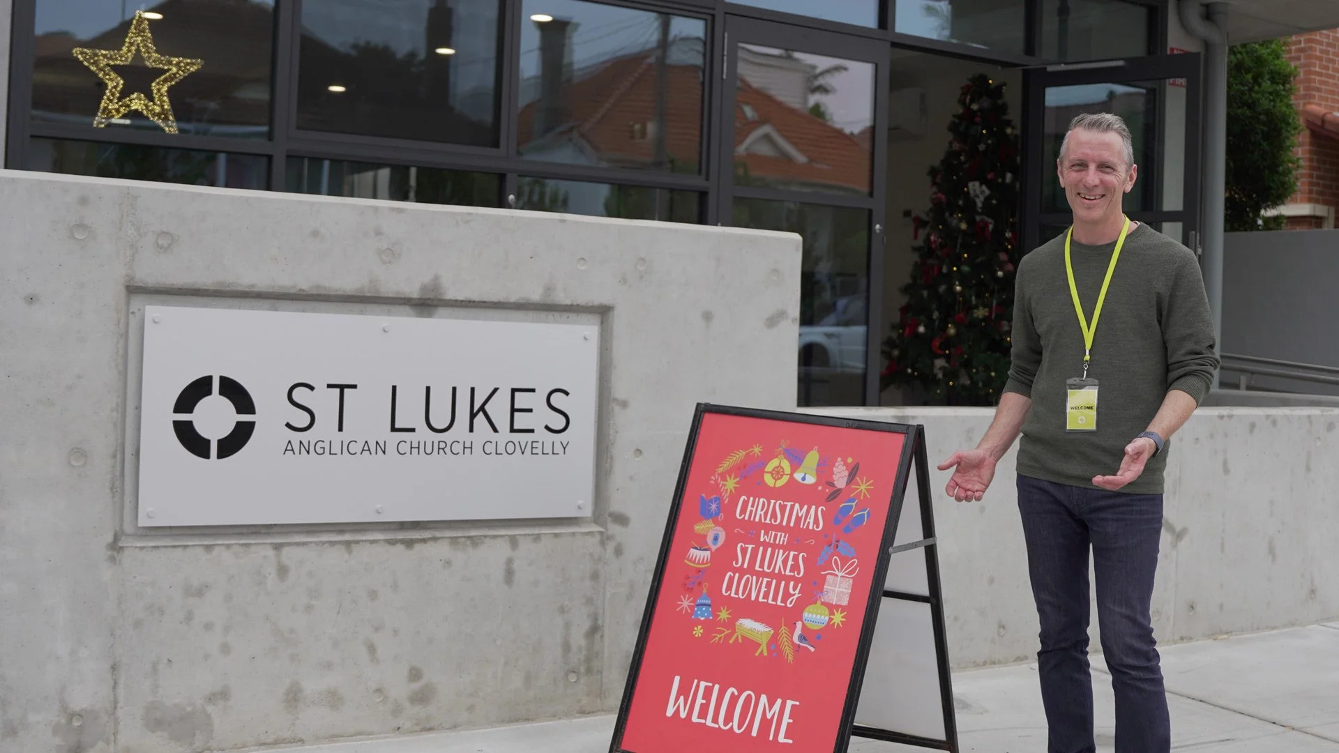 Man standing outside St Lukas Anglican Church Clovelly, smiling, gesturing with open arms, near a festive Christmas sign, with Christmas tree inside the church visible in the background.