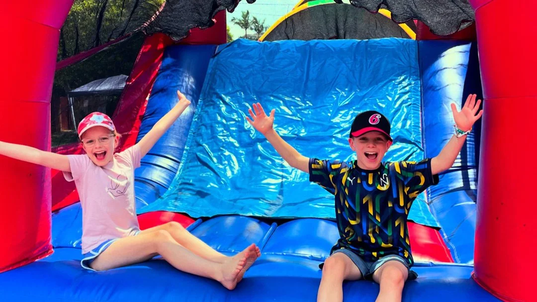 Two children on an inflatable bounce house slide, smiling with their arms raised, outdoors on a sunny day.
