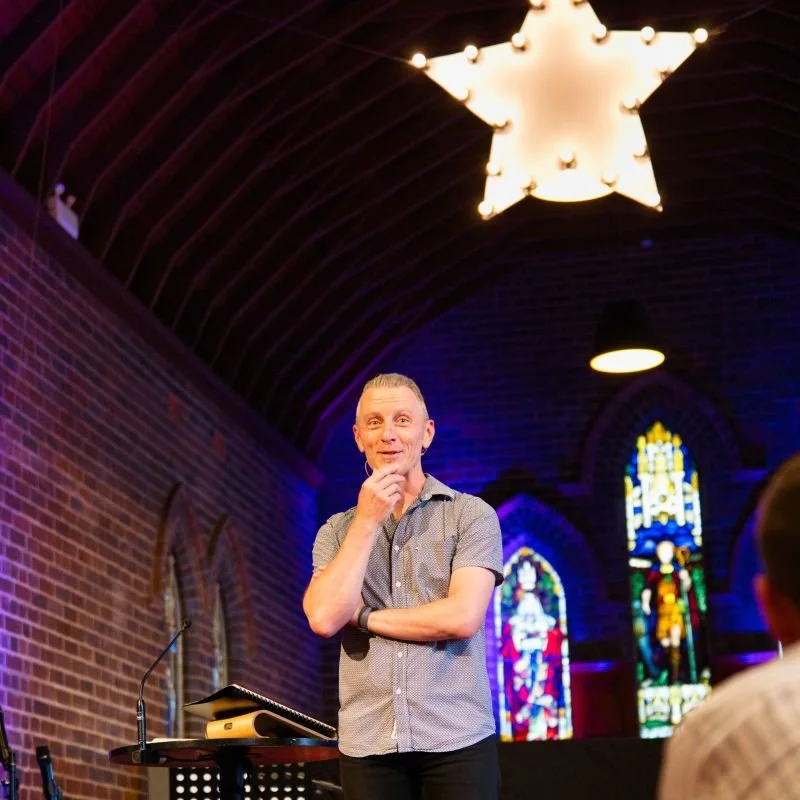 A man standing in front of two stained glass windows inside a church, facing the audience, with a large illuminated star-shaped light hanging from the wooden ceiling.