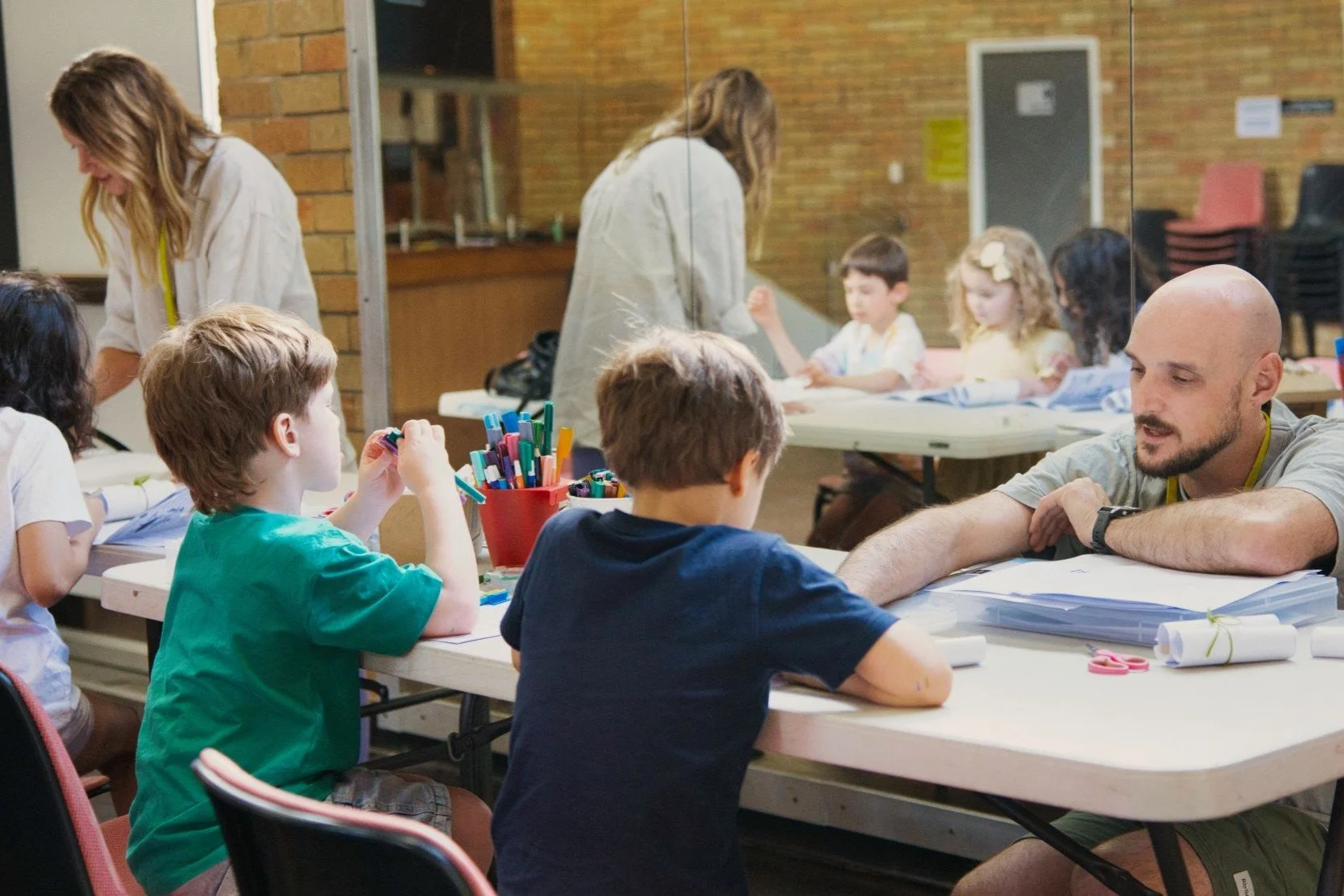 Children and adults in a classroom engaged in activities, with some children sitting at tables and adults assisting them, separated by a glass partition.