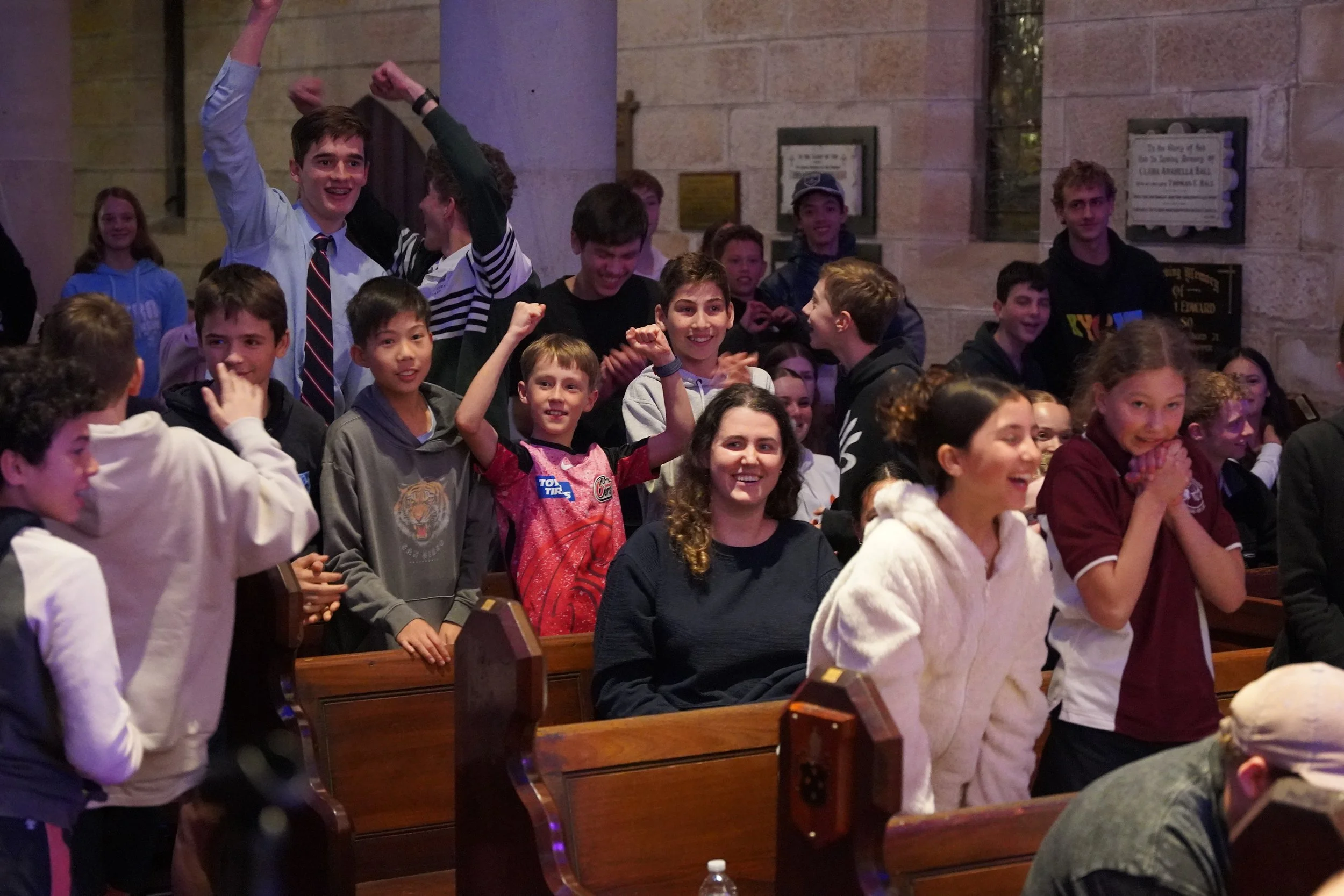 Group of children and a woman inside a church, some children are excited, laughing, and raising their fists, with wooden pews and stone walls in the background.