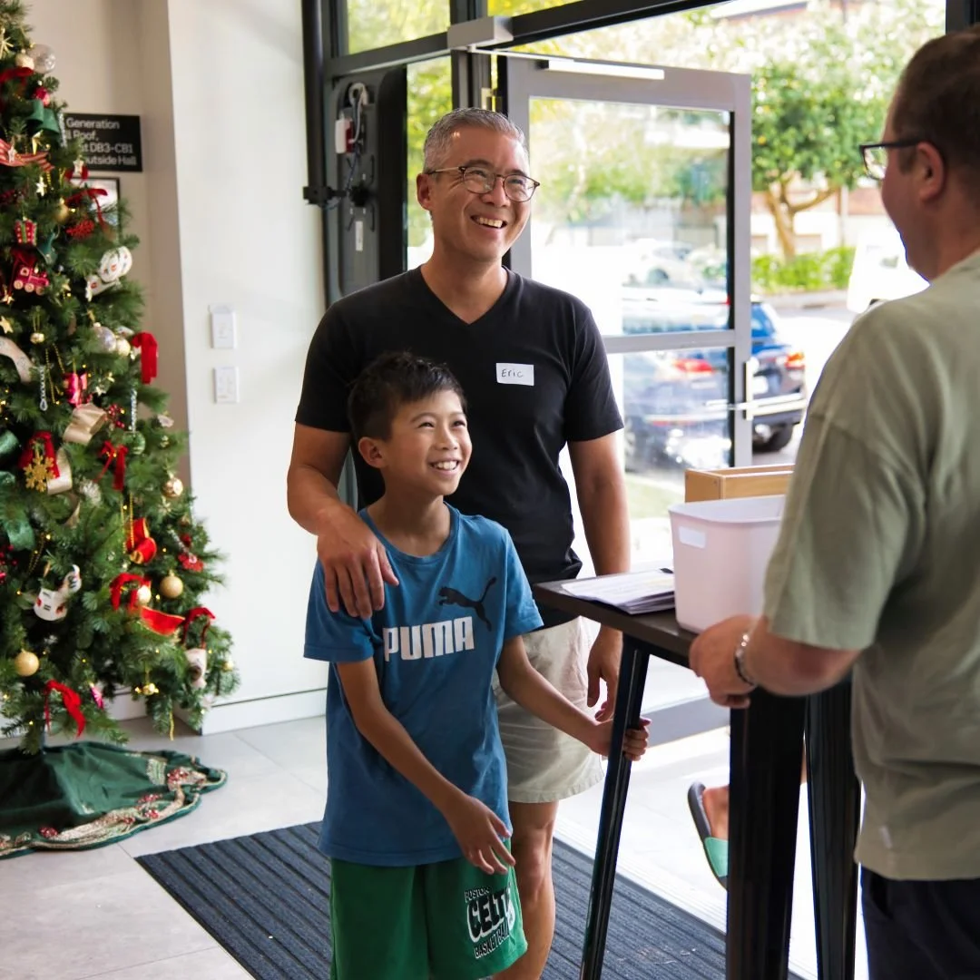 A man with glasses and a young boy are smiling and talking to a person at a reception desk. There's a decorated Christmas tree on the left side of the image, suggesting a holiday setting.