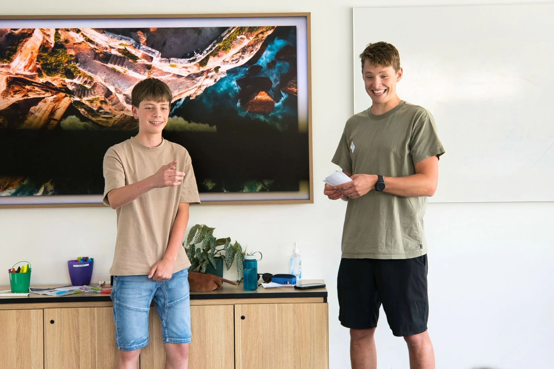 A teenage boy and an adult man standing in a classroom or office, smiling and talking. The boy is wearing a beige t-shirt and denim shorts, gesturing with his right hand. The man is wearing a gray t-shirt and black shorts, holding papers. Behind them is a large framed aerial photograph of a canyon or river landscape, on a white wall. A wooden cabinet with office supplies and a potted plant is visible below the photograph.