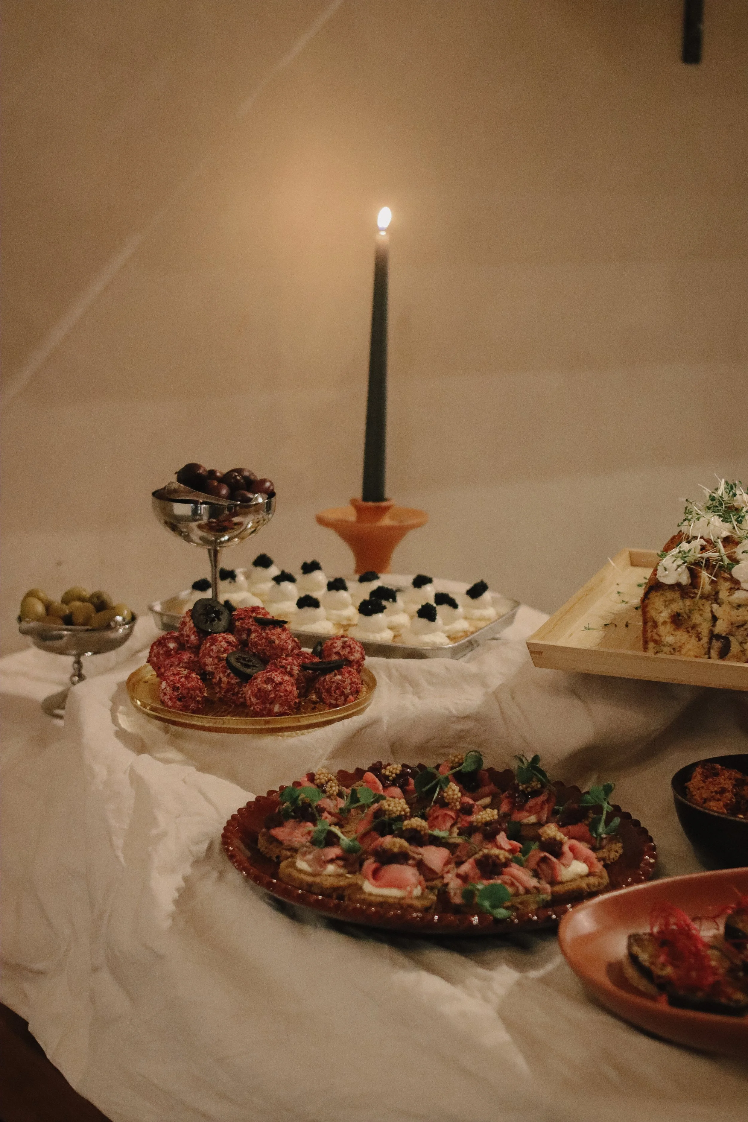 A table with various desserts, including cupcakes with white frosting and blackberries, a tray of thin crackers with pink toppings and greenery, and bowls of olives, with a lit candle in the background.