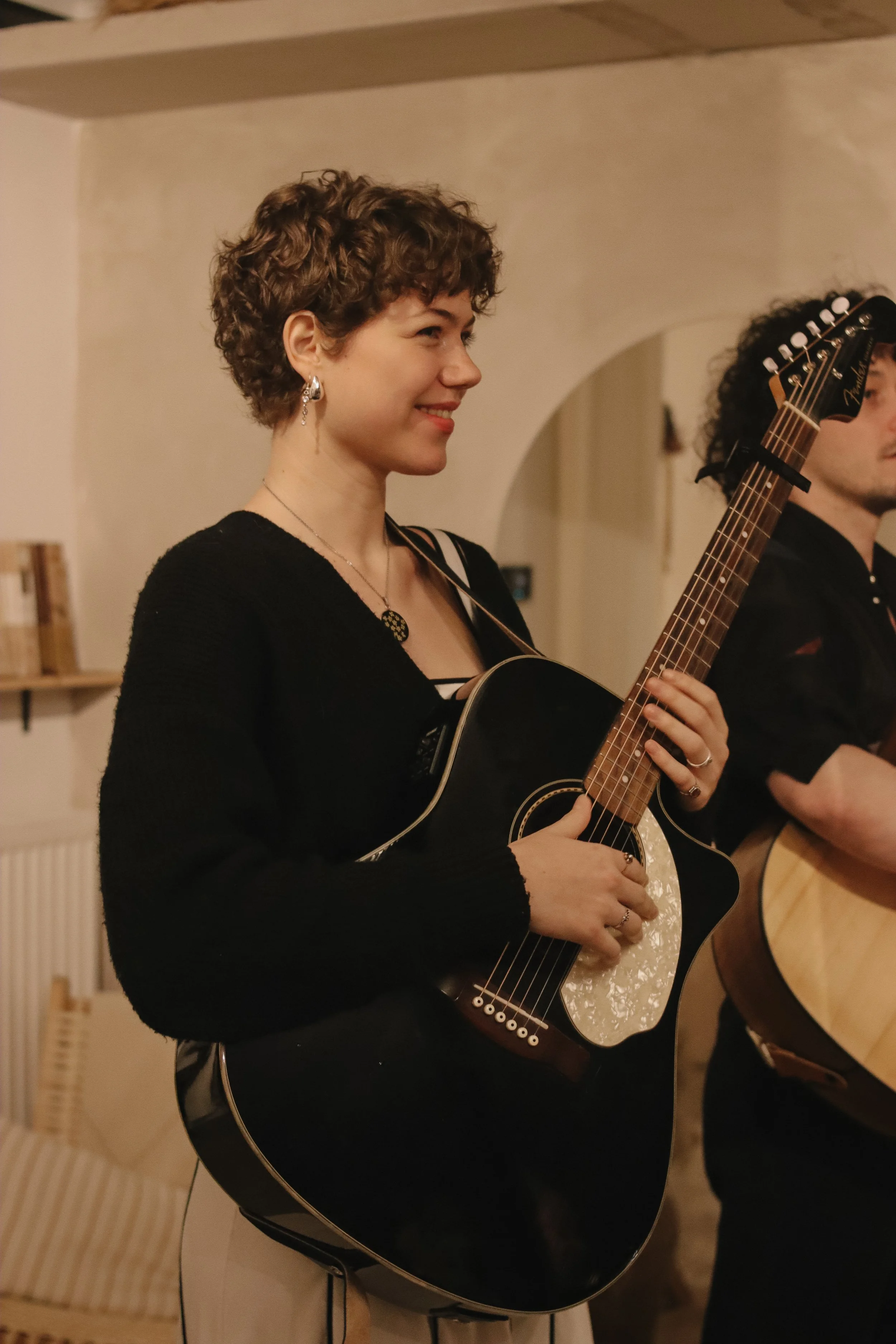 A woman with short, curly brown hair playing a black acoustic guitar in an indoor setting, smiling, with another person partially visible holding a guitar next to her.