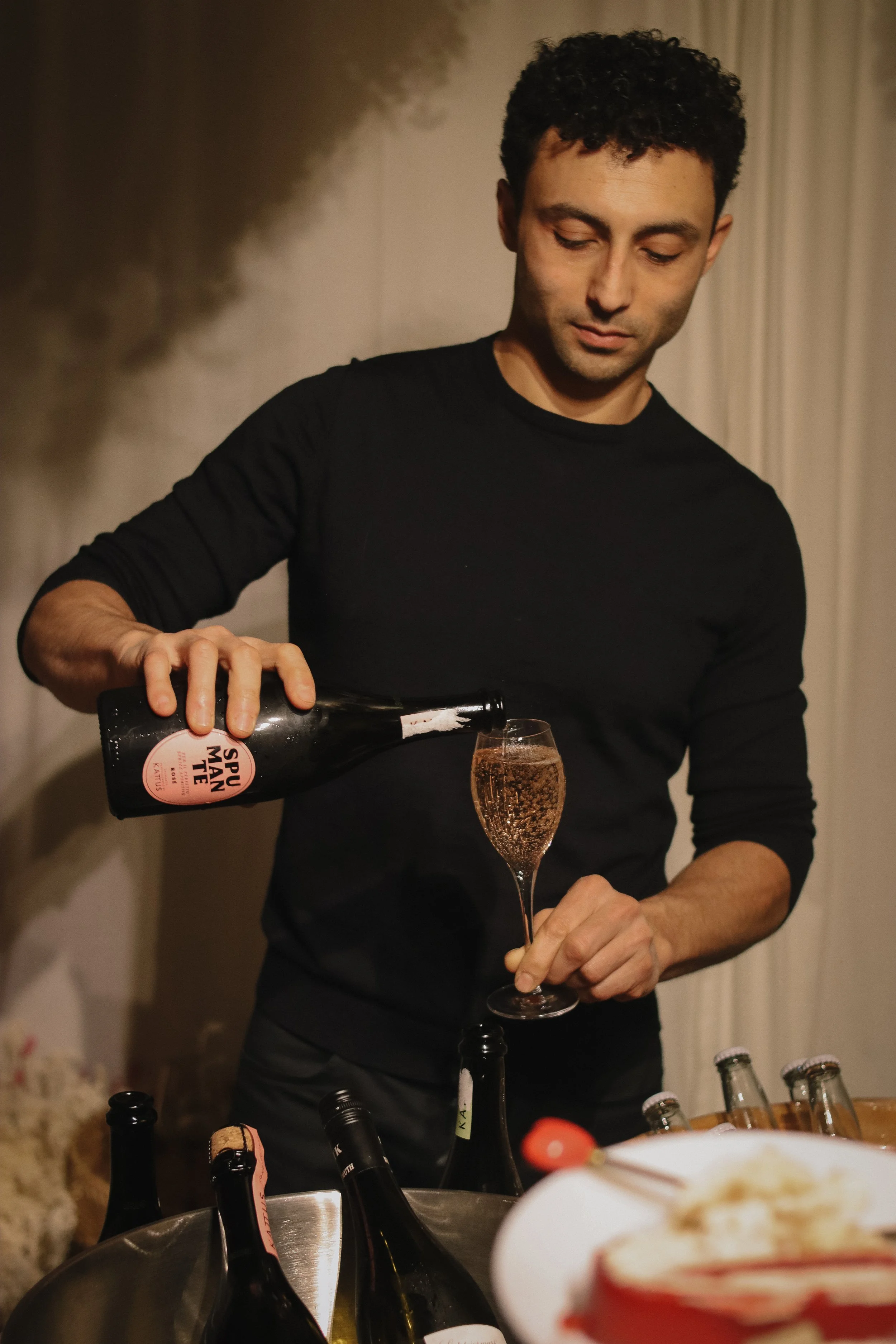 A man in a black shirt pouring sparkling rose wine into a champagne flute at a gathering.