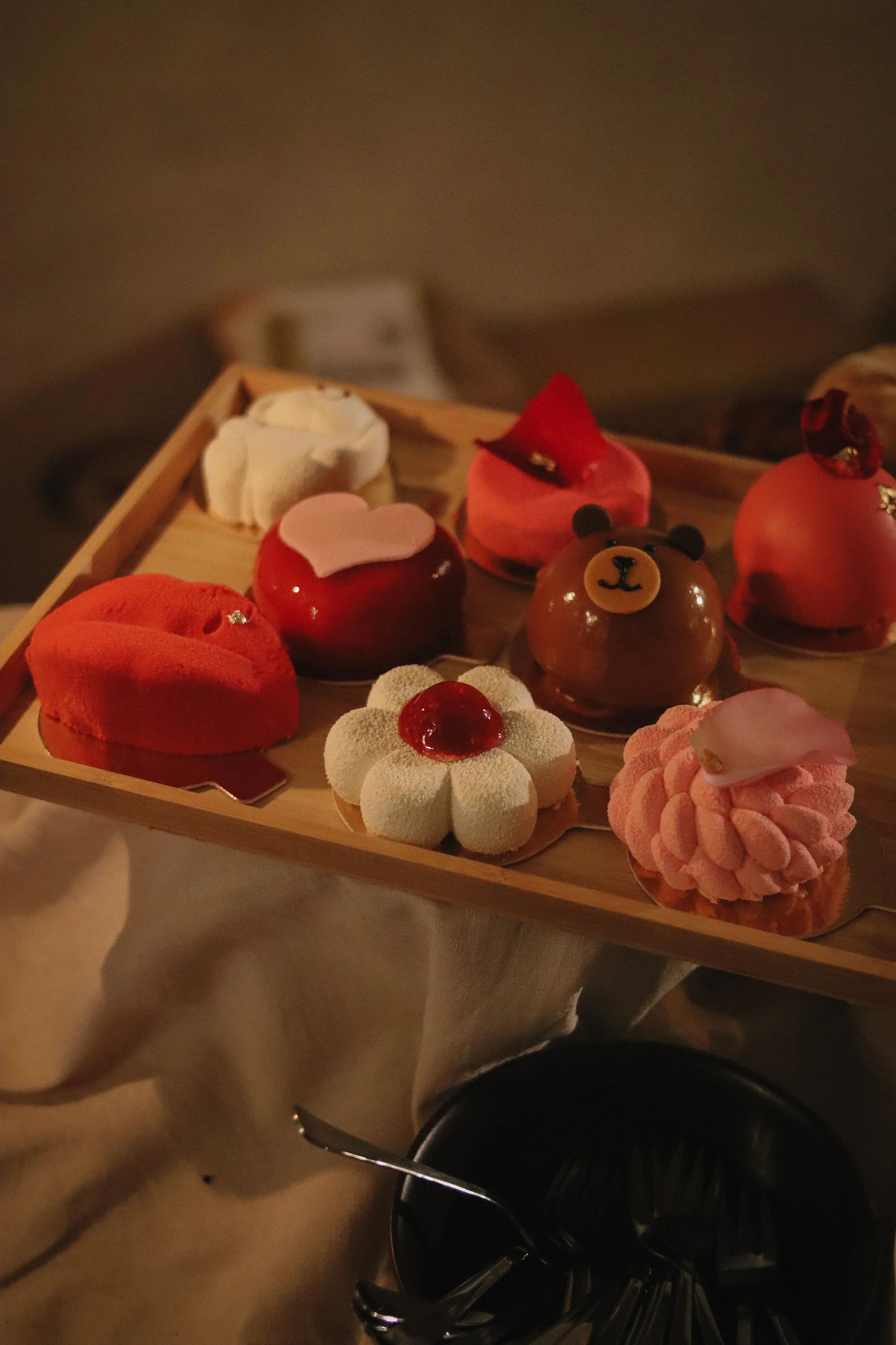 Assorted colorful pastries arranged on a wooden tray, including a bear-shaped pastry, a flower-shaped pastry with a cherry on top, and various other desserts, placed on a table with a dark background.