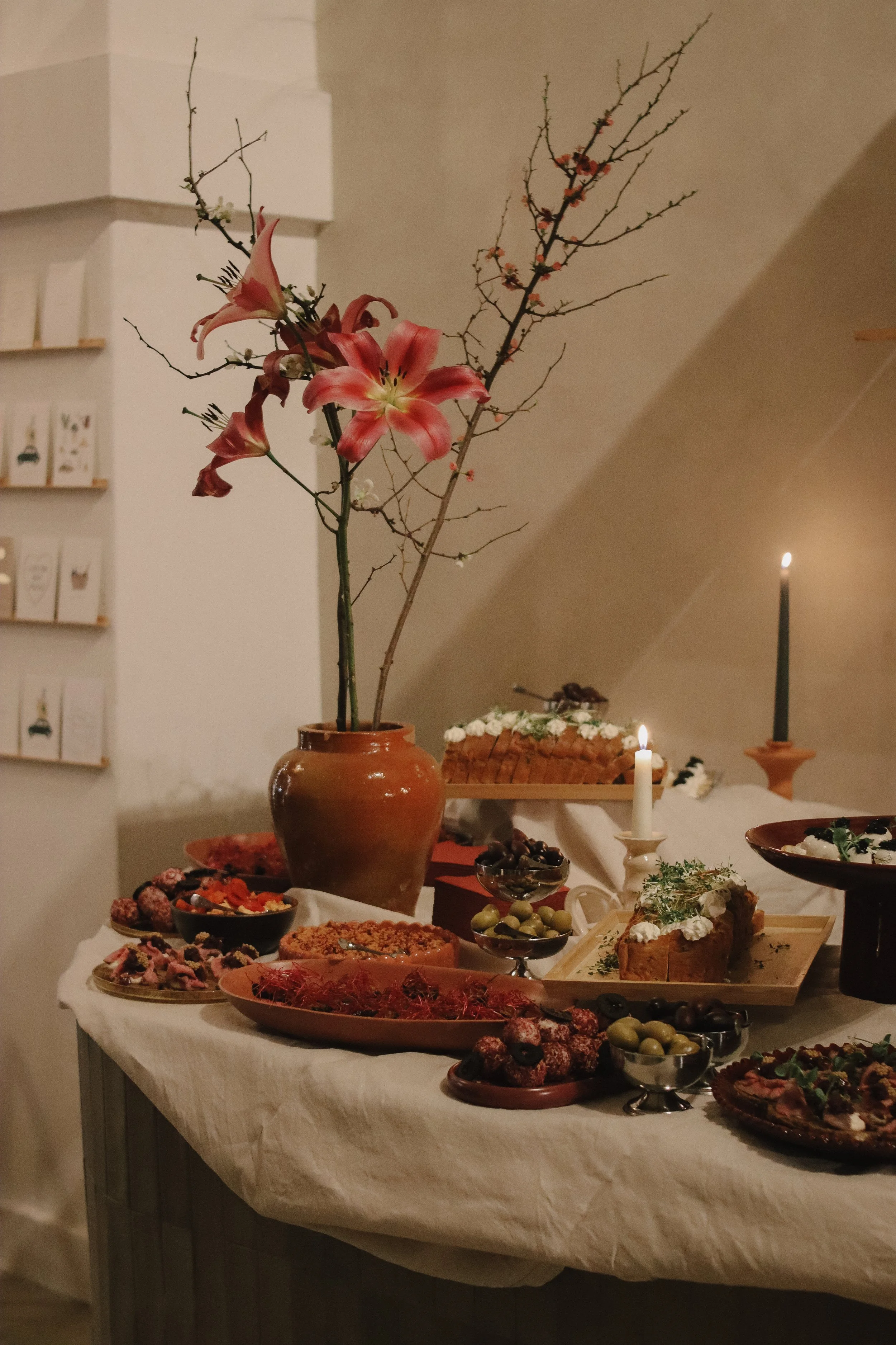 A dessert table with a large brown vase holding pink lilies and bare branches, surrounded by various pastries, fruit, and decorated cakes, with lit candles in a cozy indoor setting.