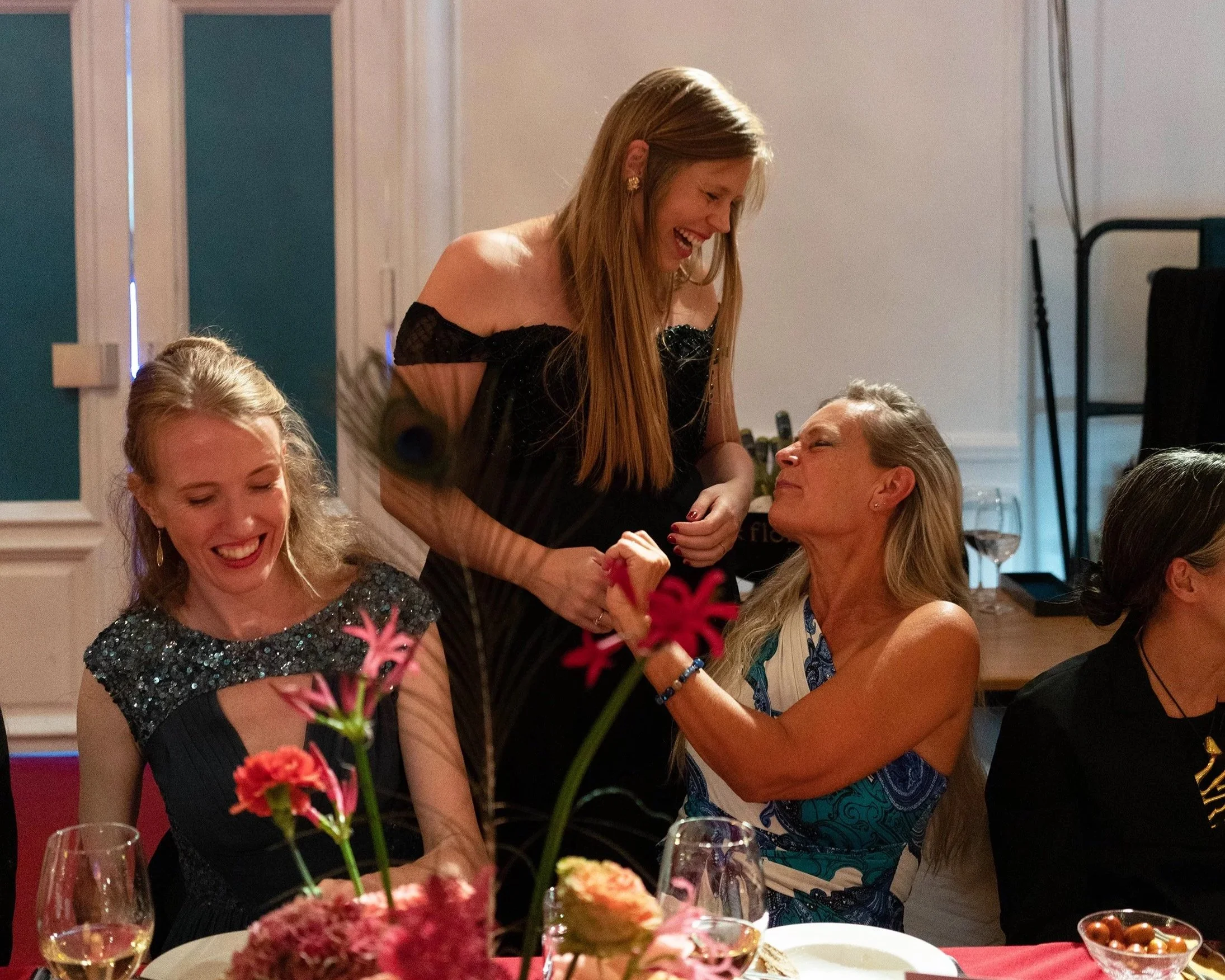 Four women at a celebration, smiling and interacting around a table with flowers, drinks, and food in a decorated room.