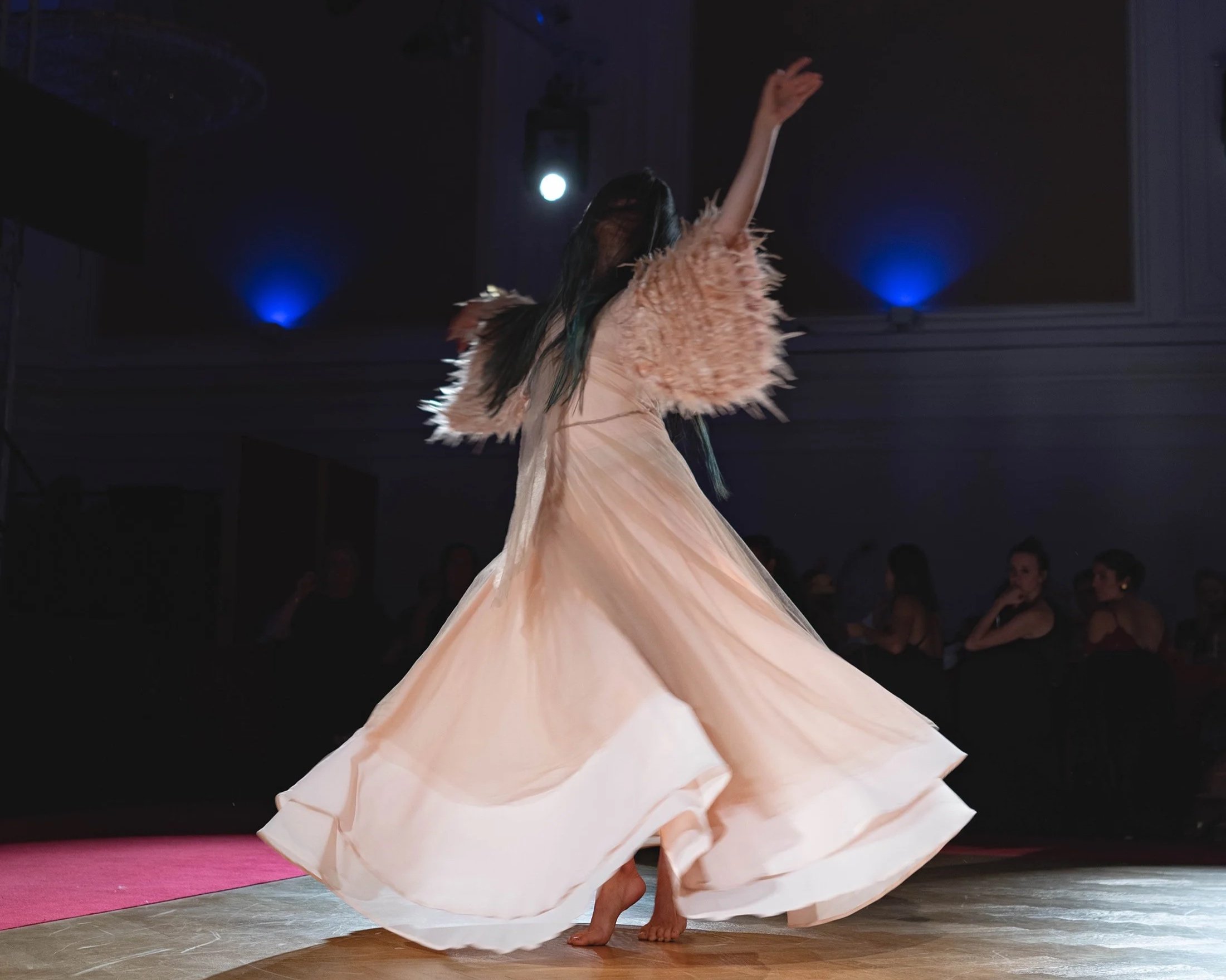 A person dancing on stage in a flowing peach dress with feathered sleeves, barefoot, in front of an audience in a dark room with blue accent lights.