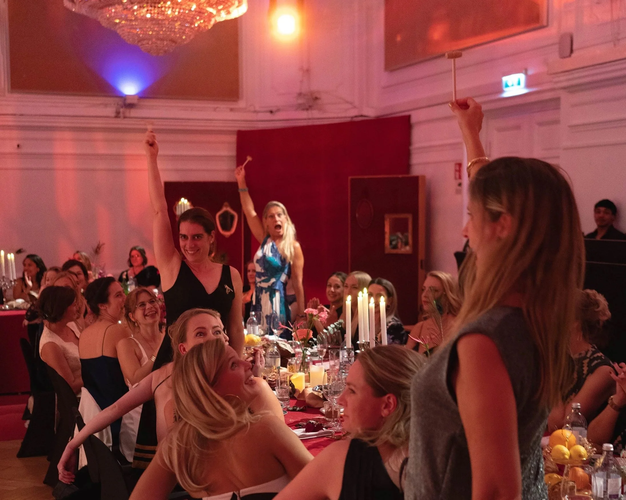 Women at a decorated dining table raising their hands with candles and flowers, in an elegant, festive event room with red walls and chandelier.