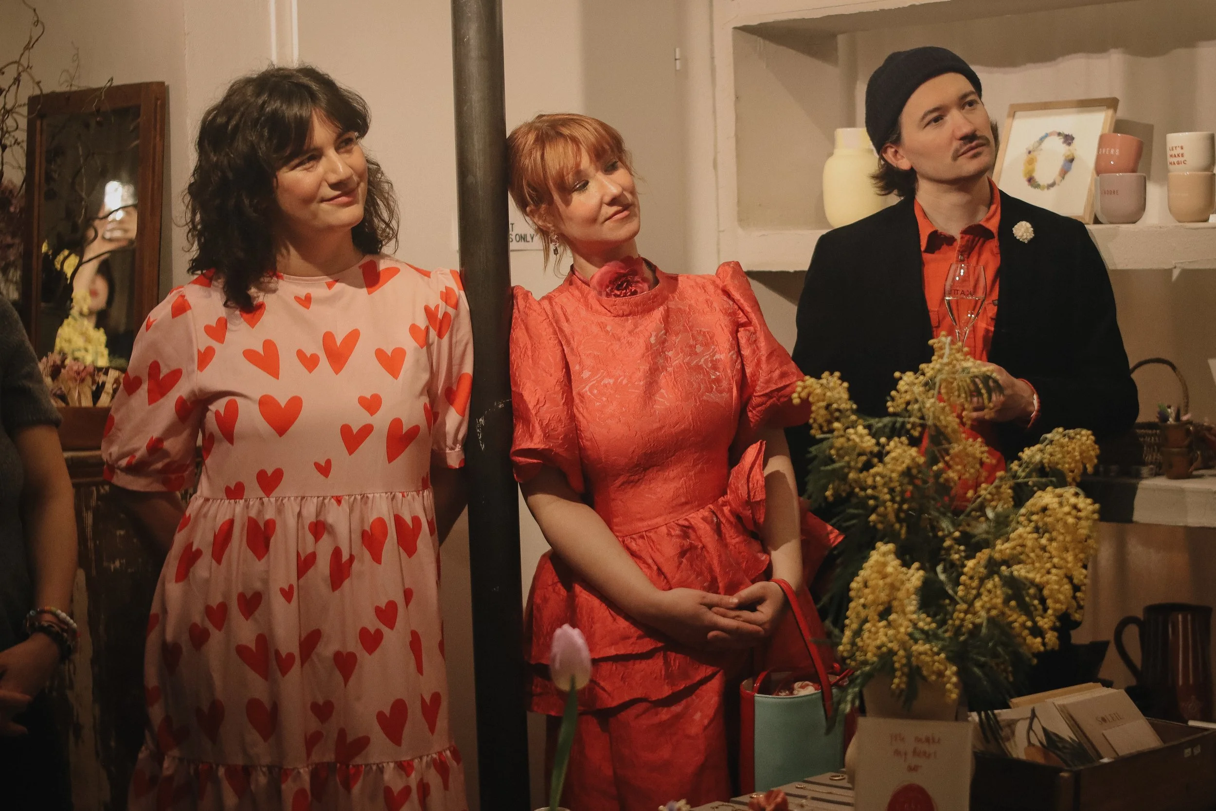 Three people in colorful outfits standing in a room with shelves and flowers, one woman in a heart-printed dress, a woman in a red textured dress, and a man in a black jacket holding a plant and a glass.