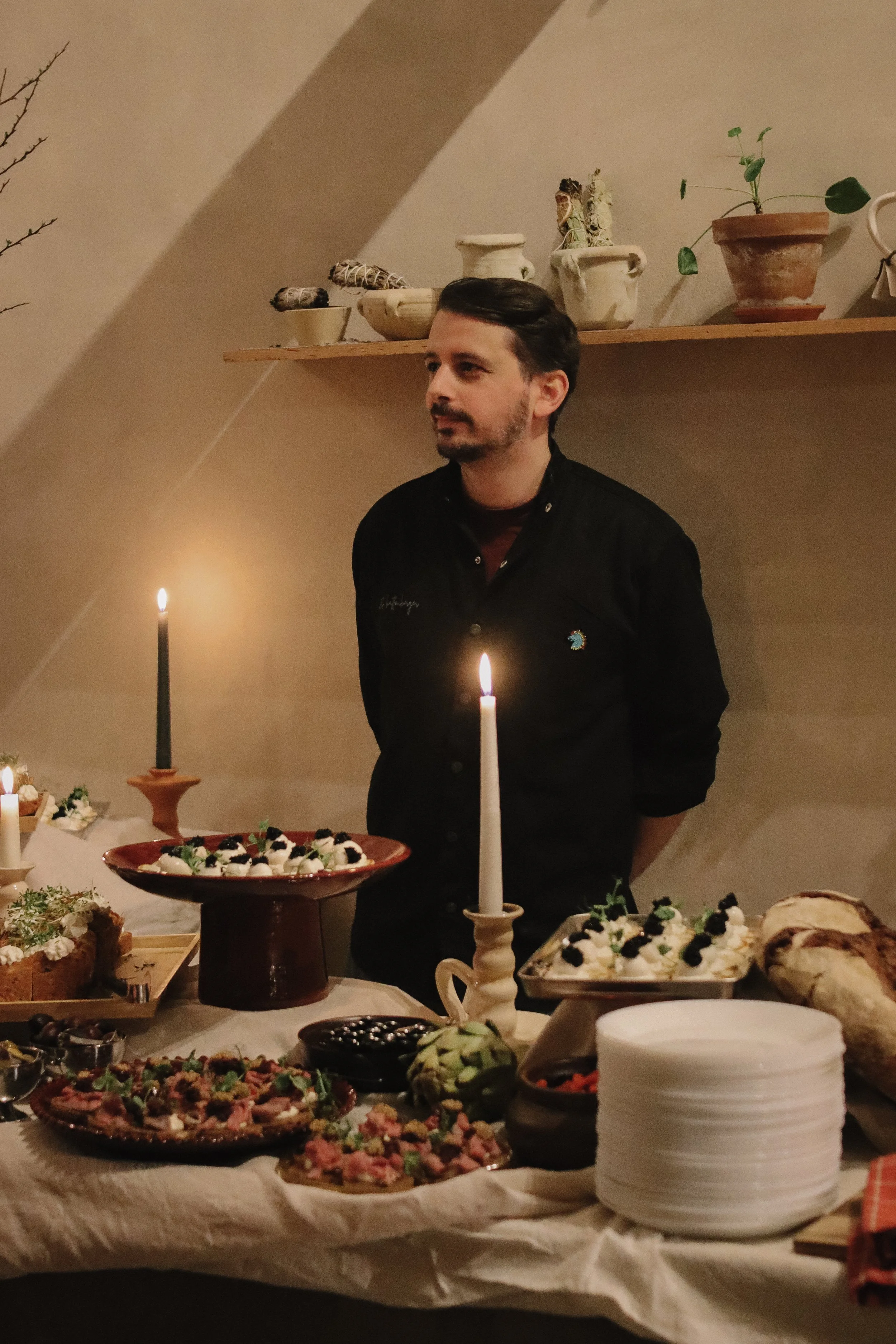 A man standing behind a table filled with various appetizers and desserts, illuminated by candlelight at a cozy gathering or celebration.