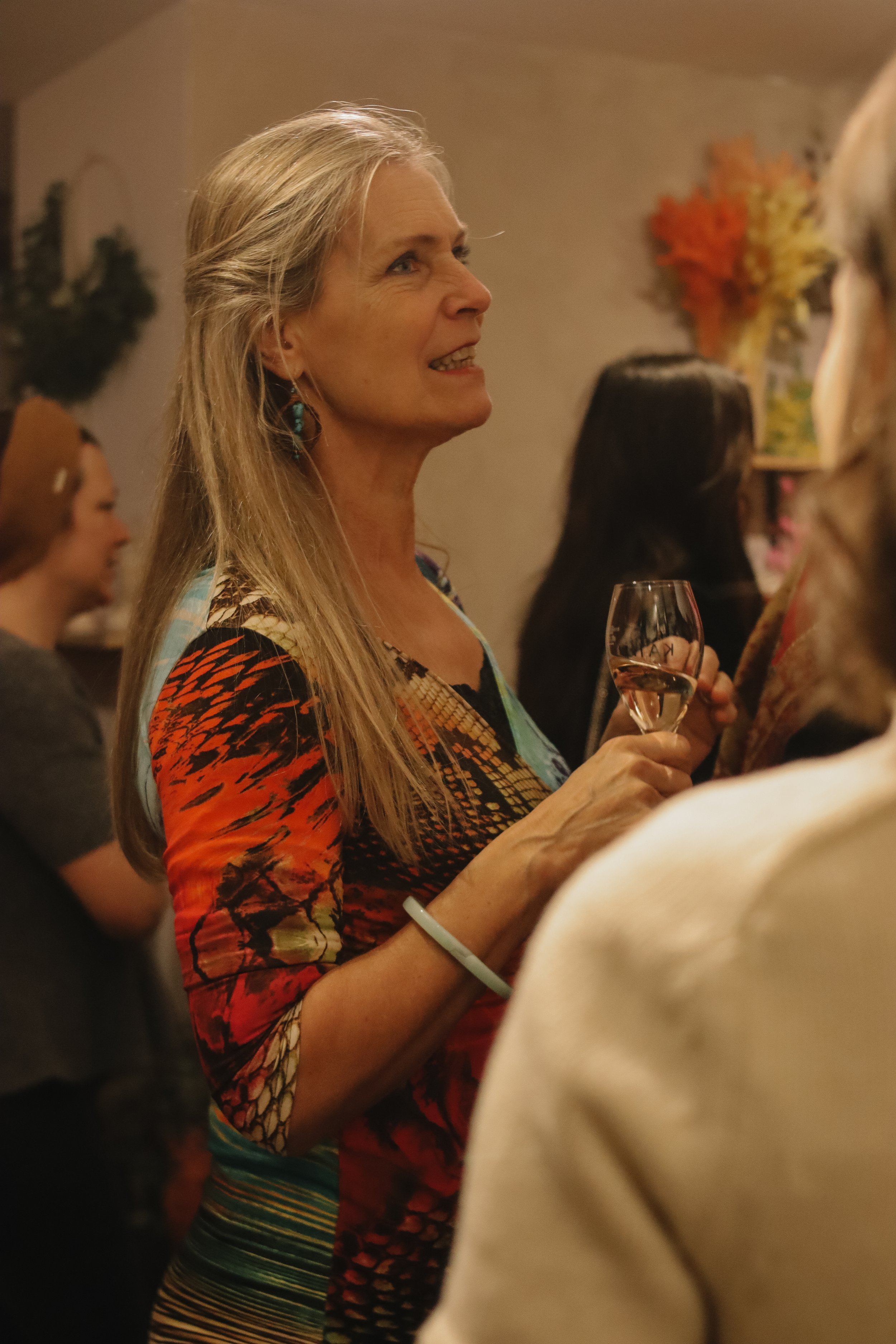 A woman with long blonde hair holding a glass of wine, engaged in conversation at a social gathering.