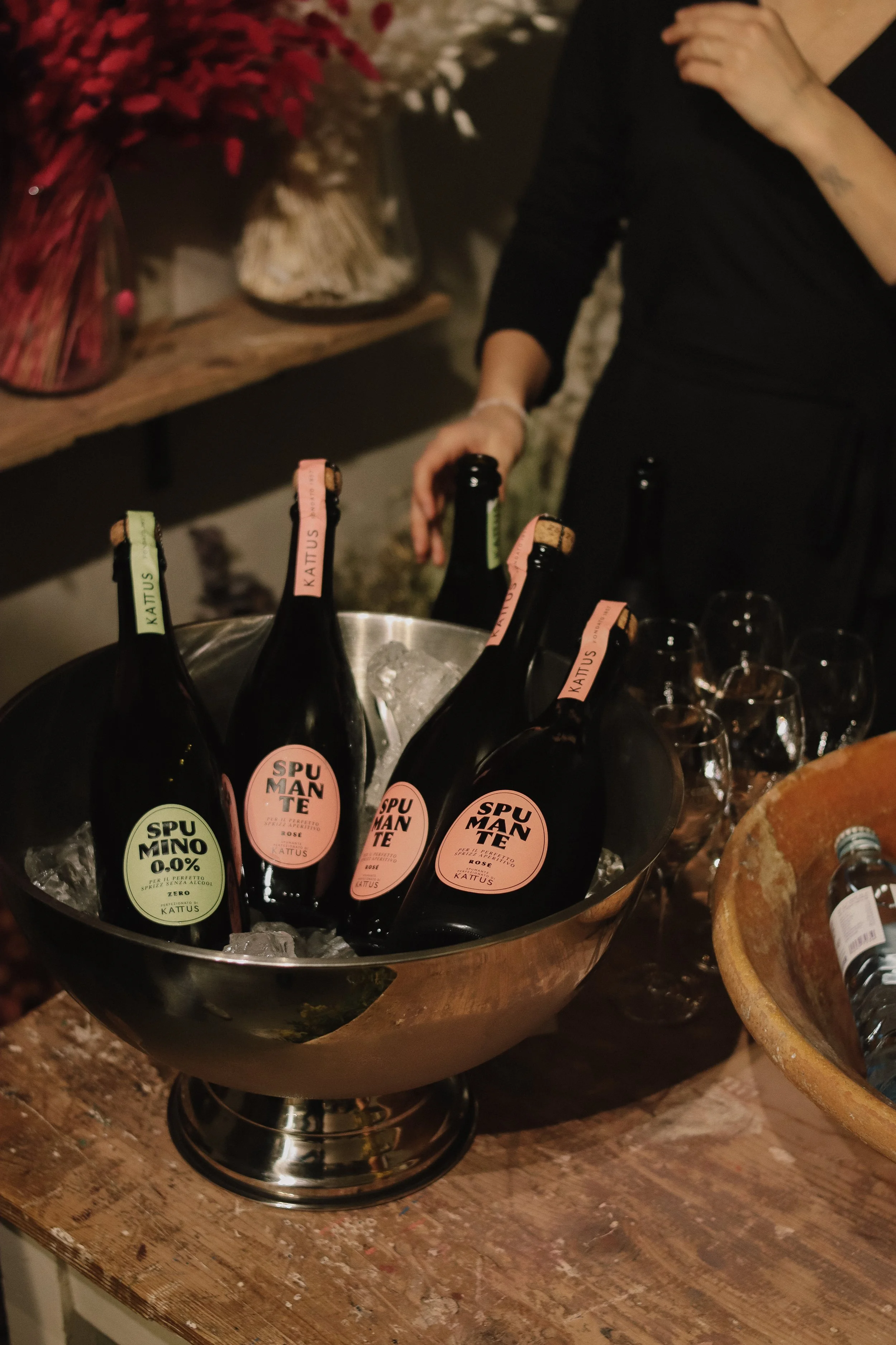 Four bottles of sparkling wine chilling in a metal bowl of ice on a wooden table, with a person reaching for one bottle and additional empty glasses nearby.