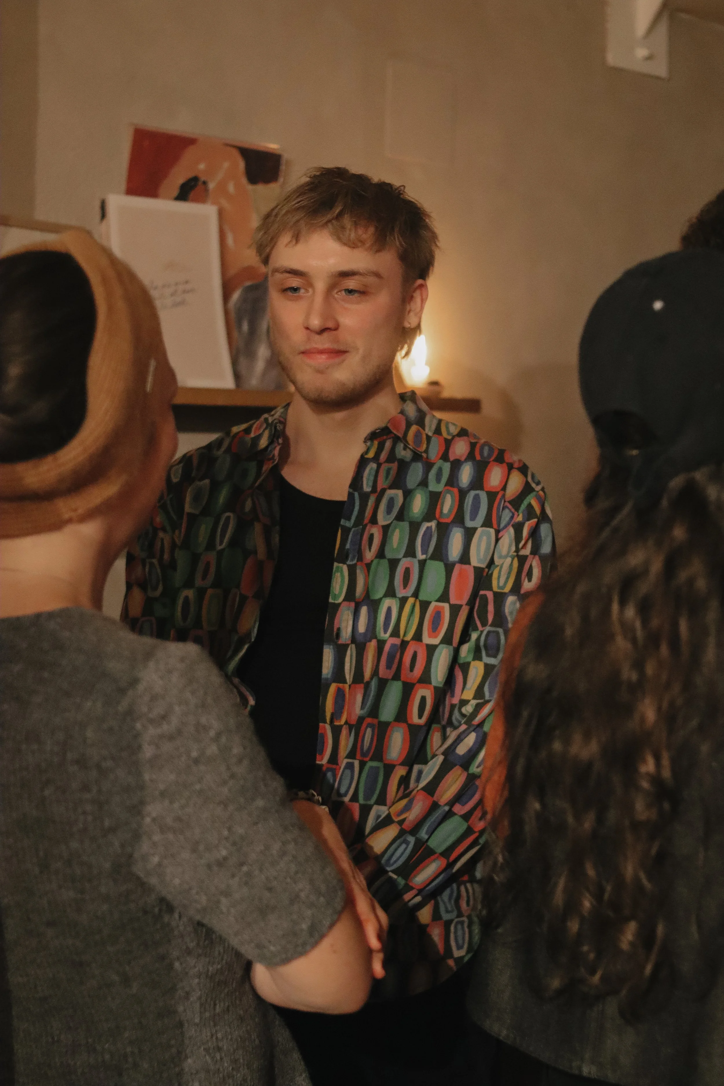 A young man with blond hair and a colorful patterned shirt chatting with two women in a cozy, warmly lit room.