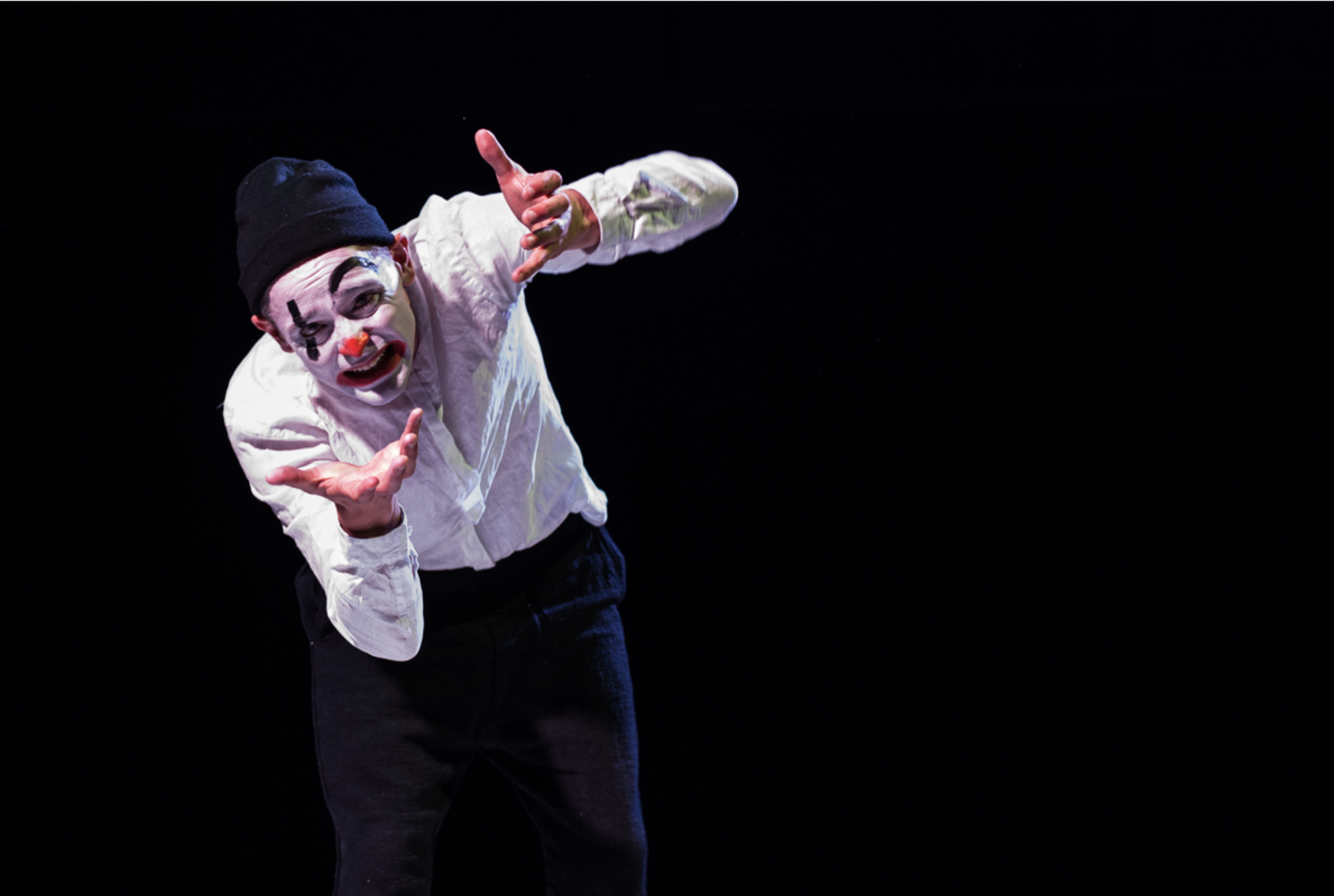 A clown with white face paint, red nose, and black eyebrows, wearing a black beanie, white shirt, and black pants, performing on a dark stage.