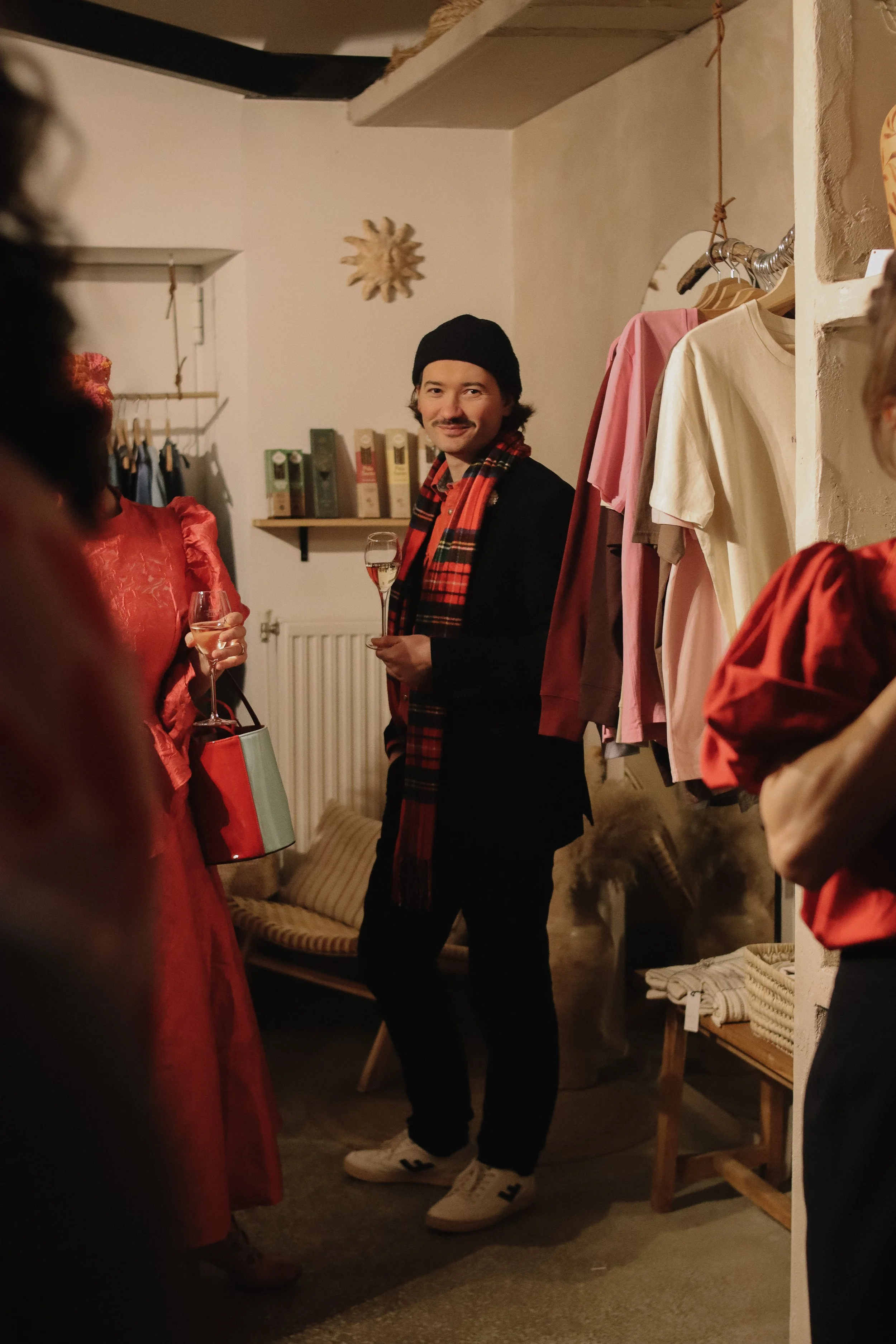 A man in a beanie and plaid scarf holding a glass of wine at a social gathering in a clothing store or boutique.