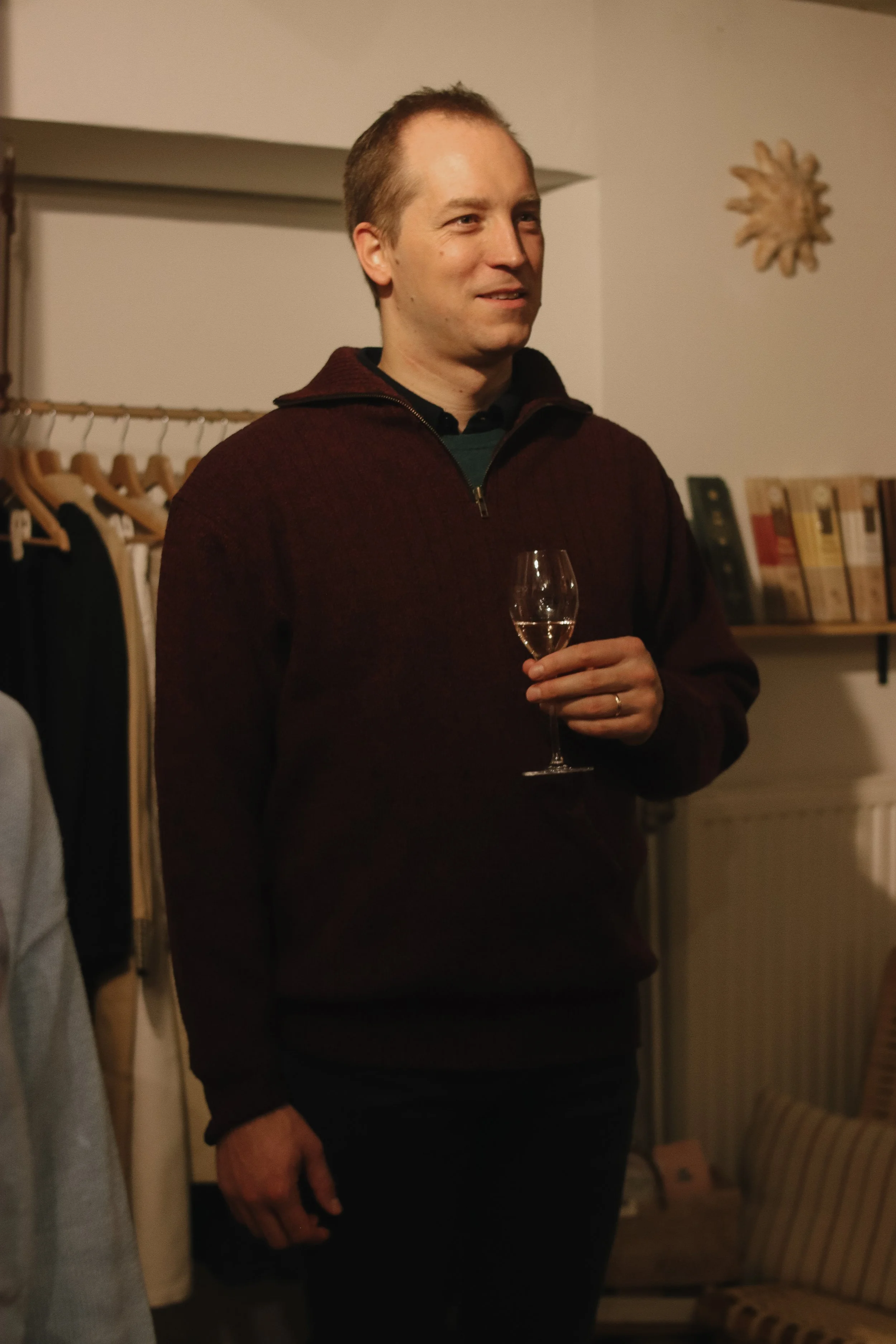 A man holding a wine glass, smiling, standing indoors with bookshelves and a decorative wall hanging in the background.