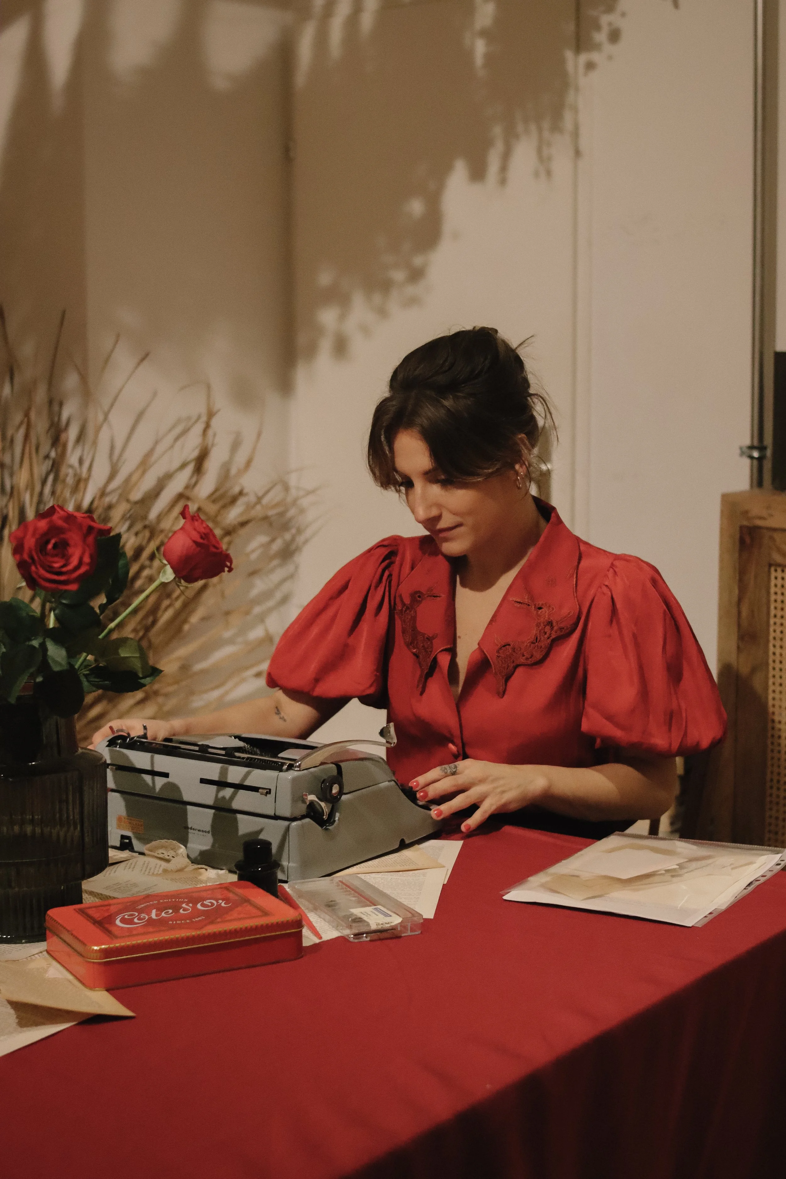 A woman in a red blouse sitting at a table with a vintage typewriter, surrounded by books, flowers, and papers, in a cozy indoor setting.