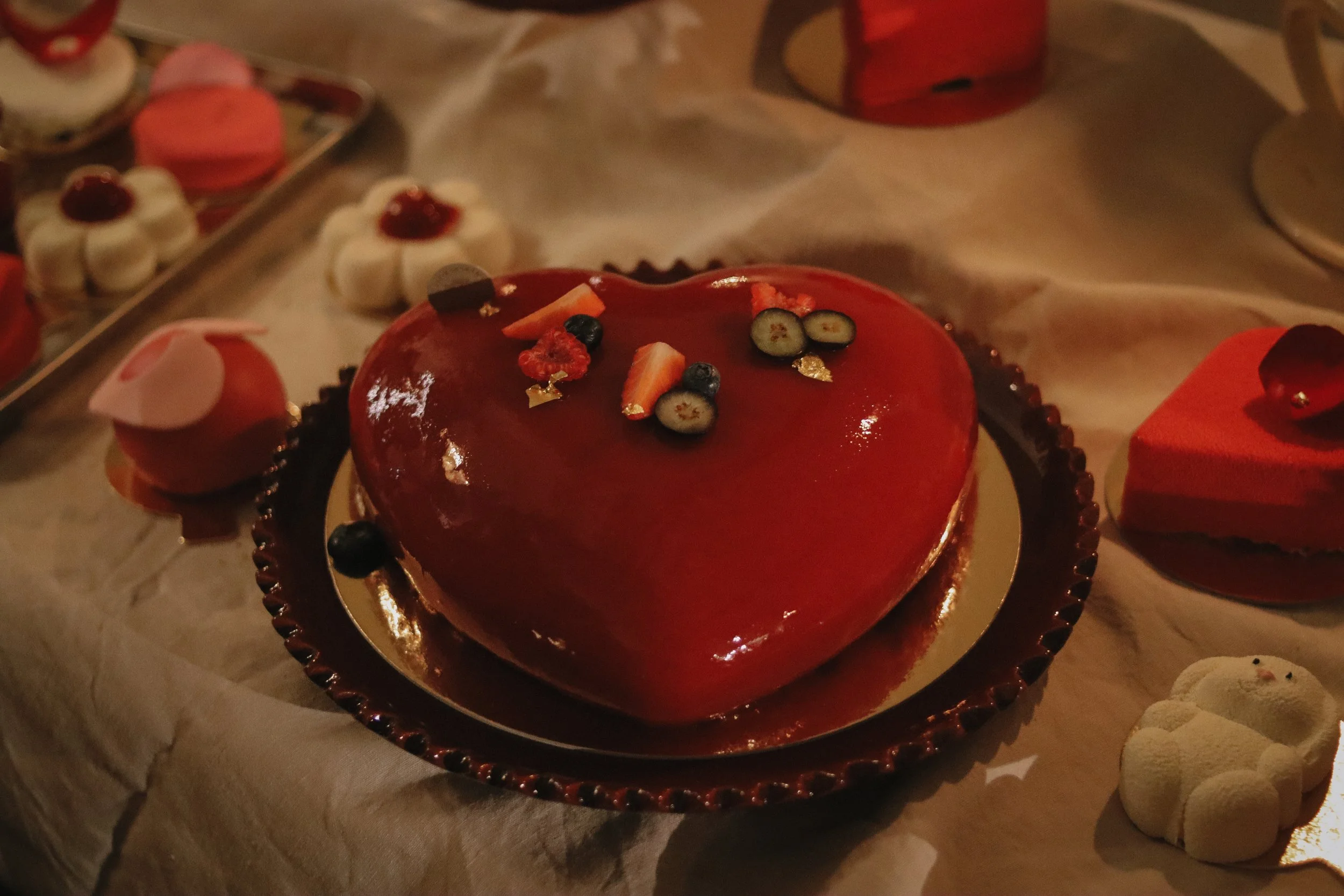 A heart-shaped glossy red cake topped with small pieces of fruit, including blueberries, strawberries, and orange slices, on a round gold and black plate. Surrounding the cake are various smaller desserts on a table with a cream-colored tablecloth, s