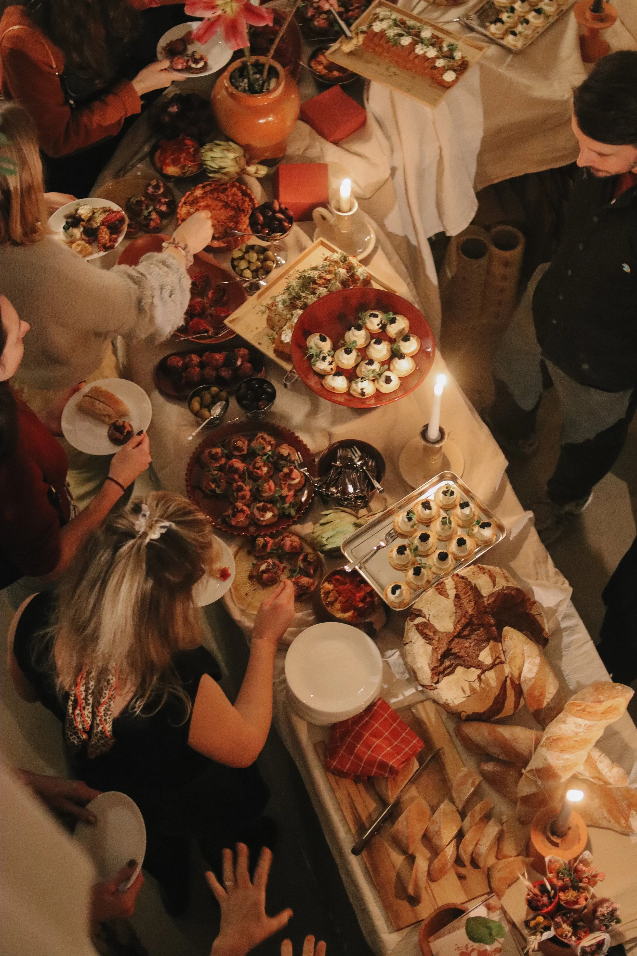 A Thanksgiving or holiday feast with various desserts, bread, and snacks on a long table, including cupcakes, rolls, pies, and fruit, illuminated by candles and surrounded by guests.