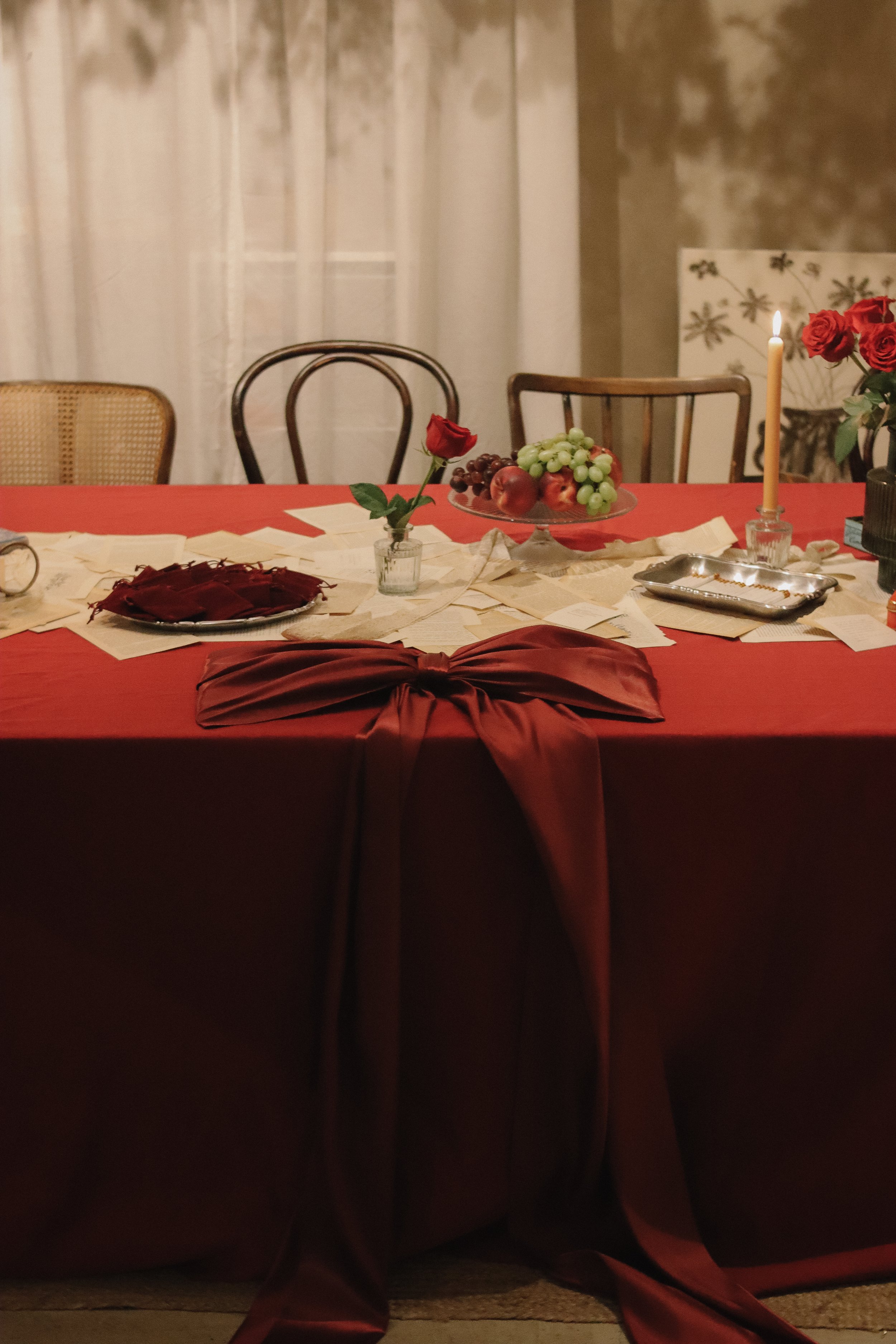 A dining table with a red tablecloth and a large red bow at the front, decorated with candles, grapes, apples, notes, and a single red rose in a vase.