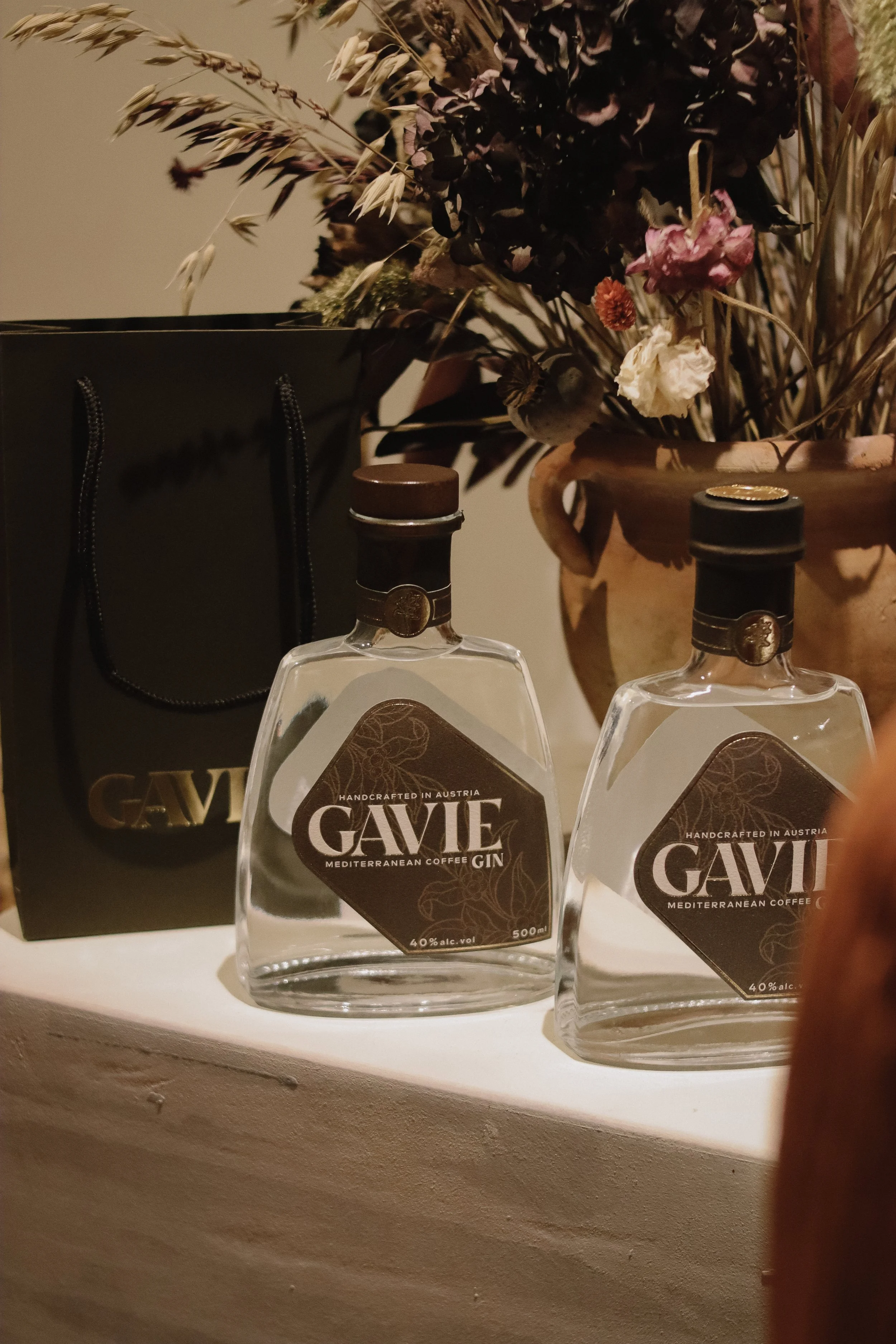 Two bottles of GAVIE Mediterranean coffee gin placed on a white surface, with a black GAVIE shopping bag and a large flower arrangement in the background.