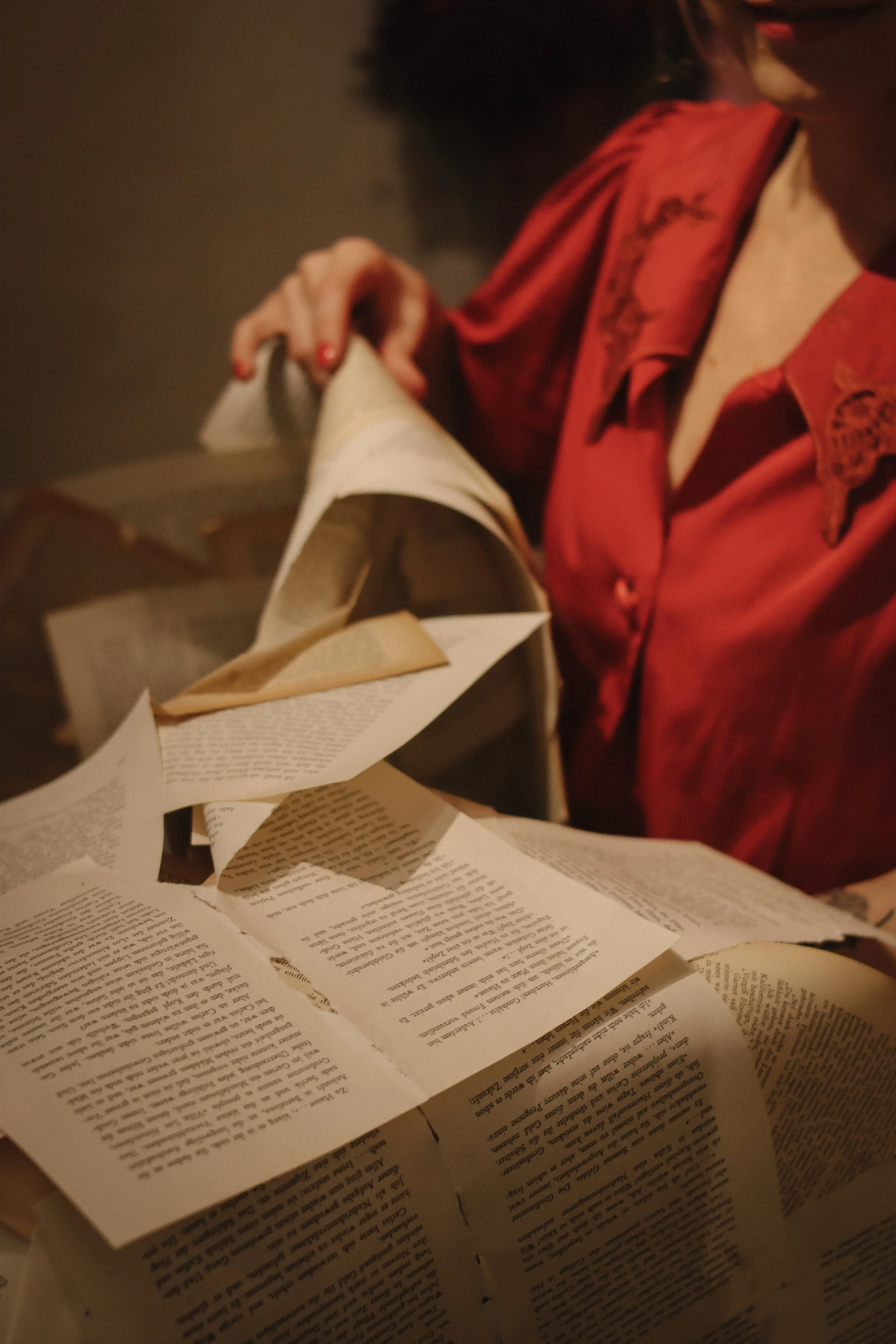 A person wearing a red shirt with lace details on the shoulders is turning the pages of a newspaper with many scattered pages on a surface.