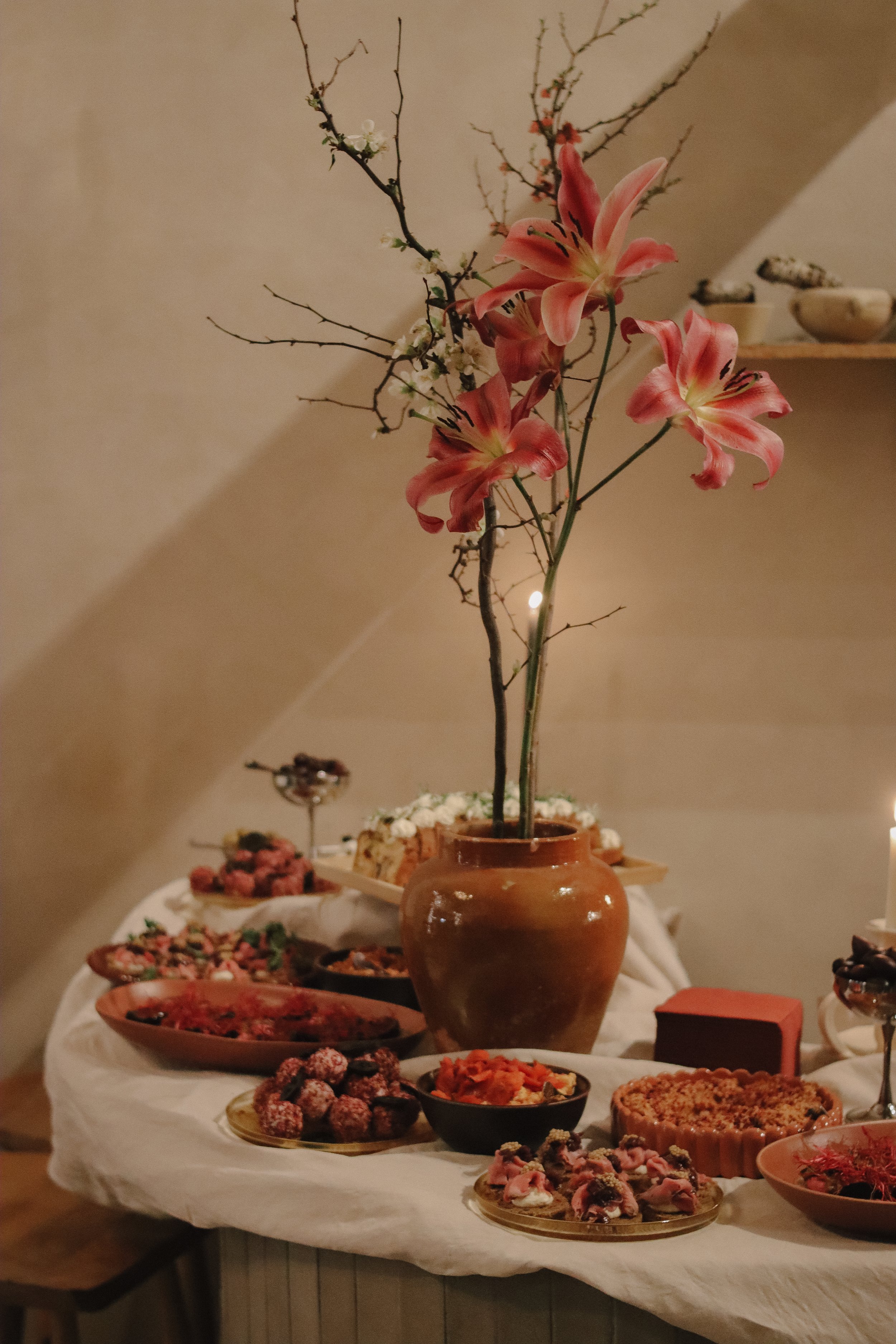 A table with various desserts and a large flower arrangement in a brown vase, featuring pink lilies and smaller branches, in a cozy indoor setting.