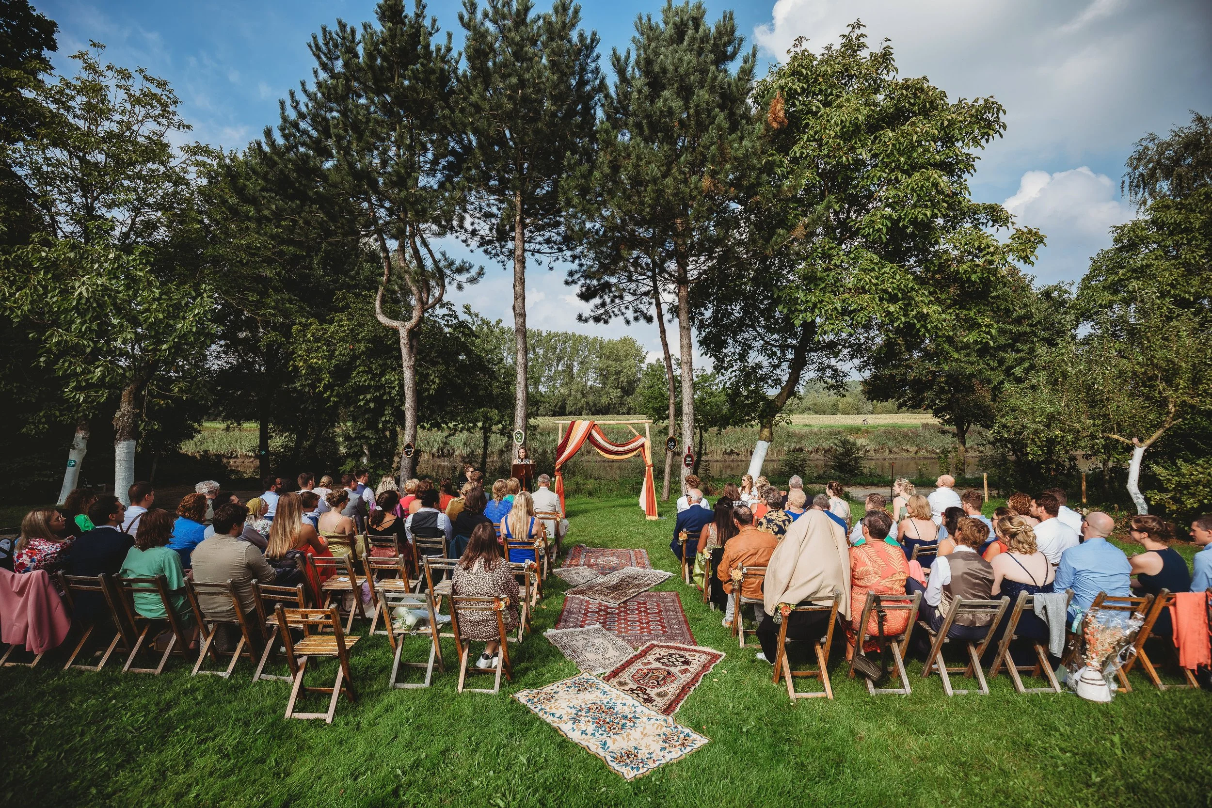 An outdoor wedding ceremony with guests seated on wooden chairs on green grass. A decorated arch is at the front, under tall trees in a natural setting with a field and trees in the background. The sky is partly cloudy.