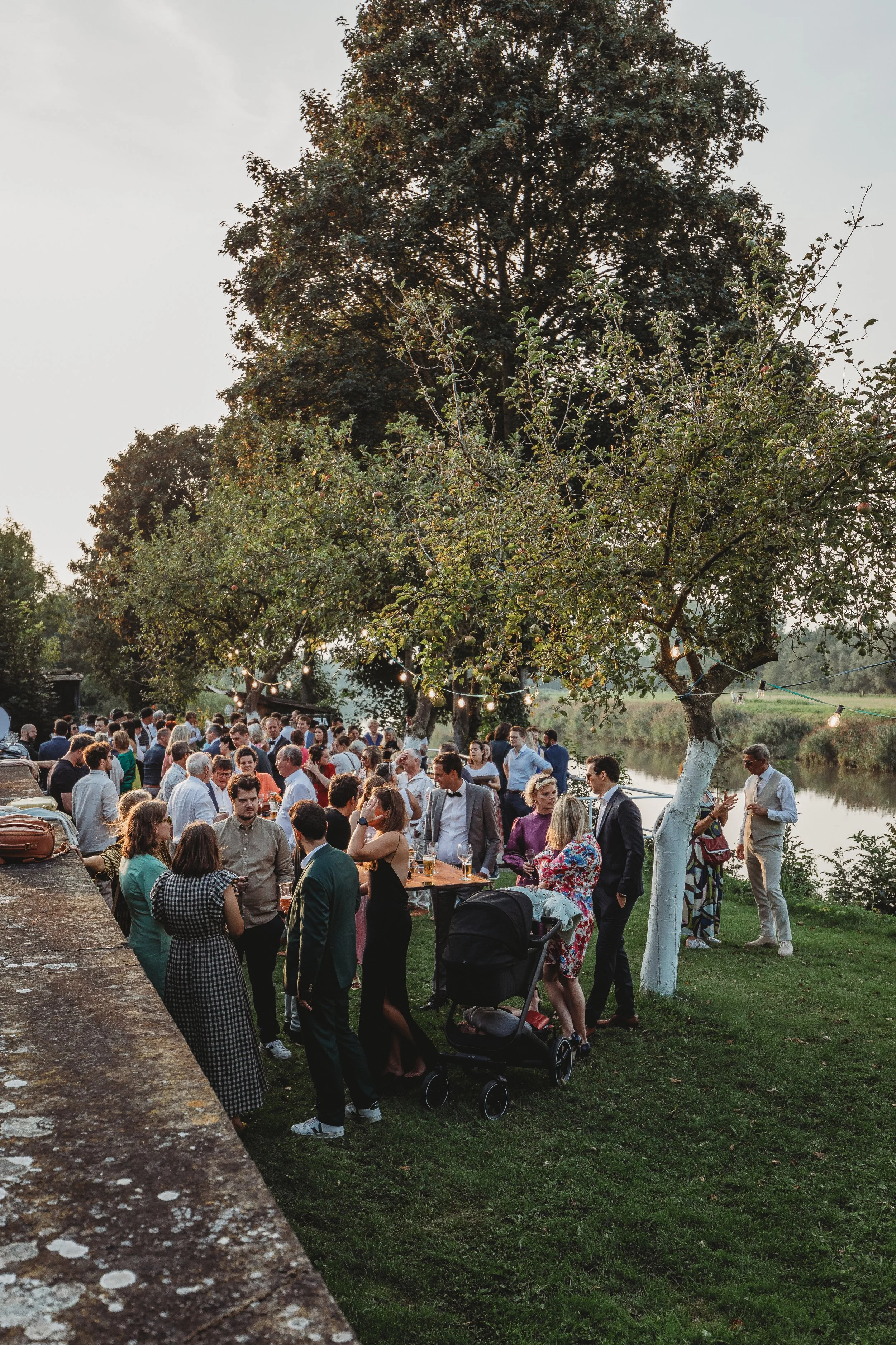 Group of people gathered outdoors on a grassy area near a river, socializing and standing under string lights, with some sitting at tables, during a daytime event.